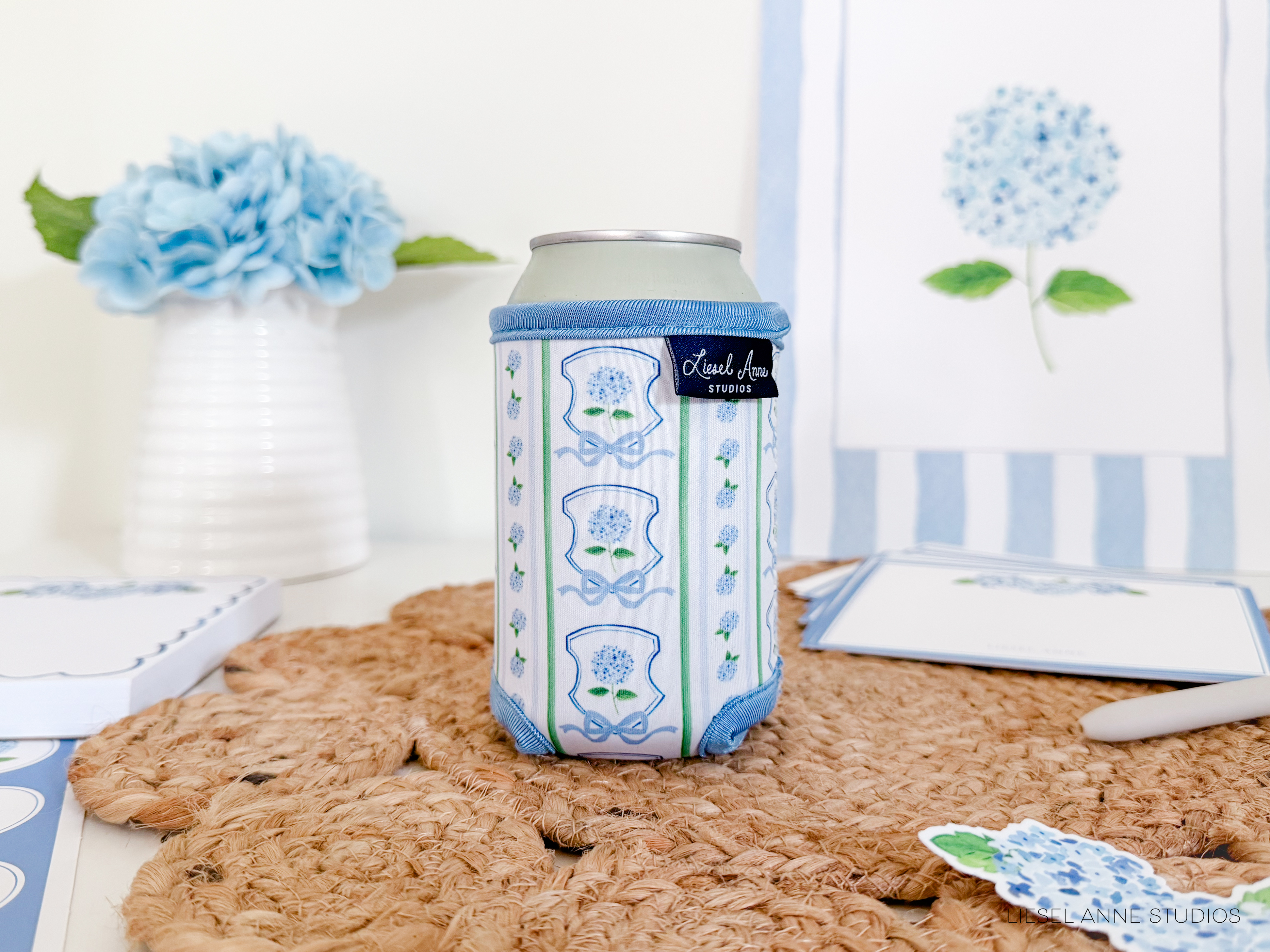 a can of soda with a blue and white floral pattern, placed on a woven placemat. In the background, there is a vase with blue flowers, and some cards and stationery items are also visible.