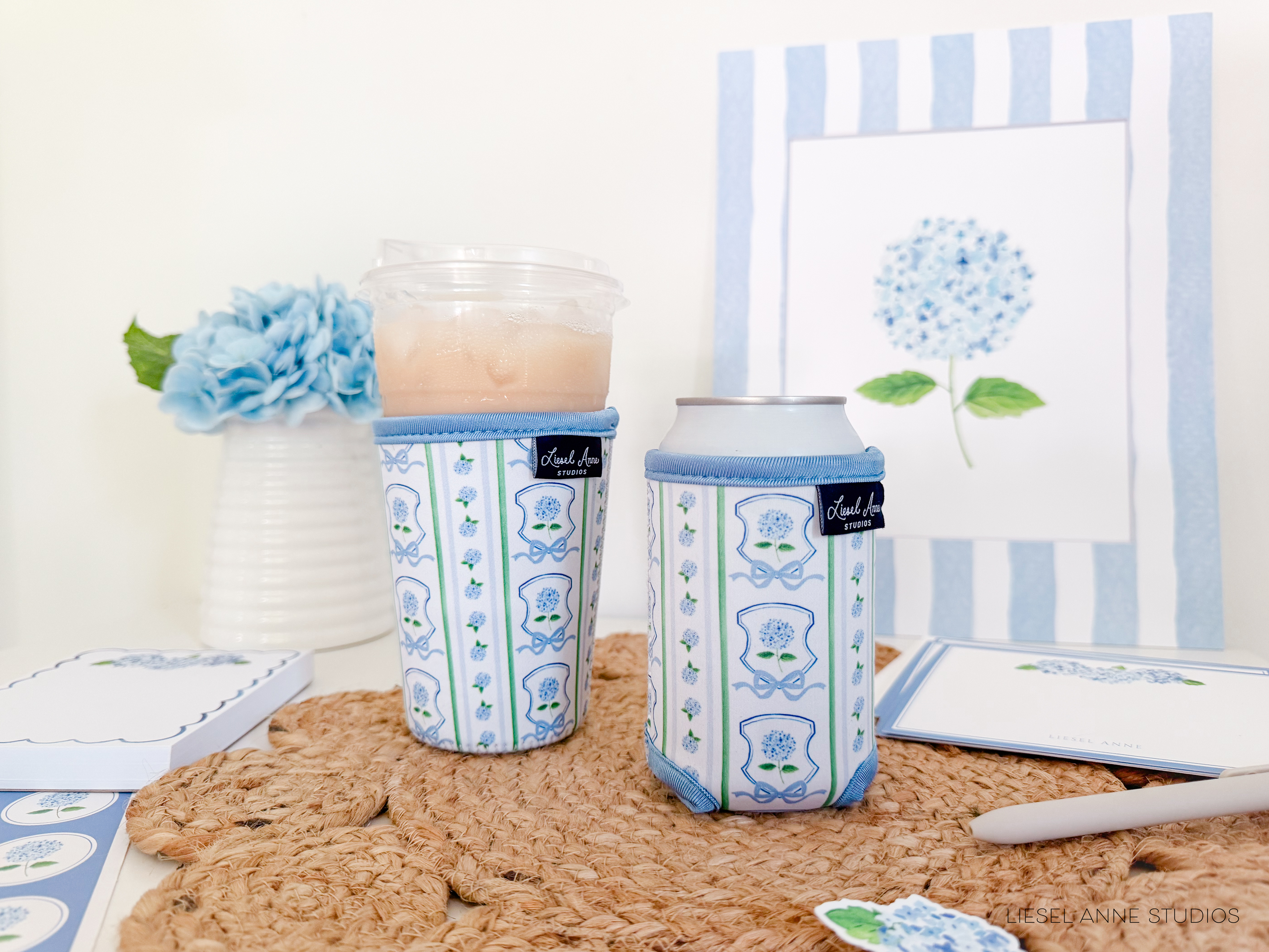a cozy and charming scene with a white vase holding a beautiful blue hydrangea flower, a white cup with a blue and white floral pattern, and a white can with a blue floral design. The objects are arranged on a woven placemat, creating a visually appealing and harmonious composition.