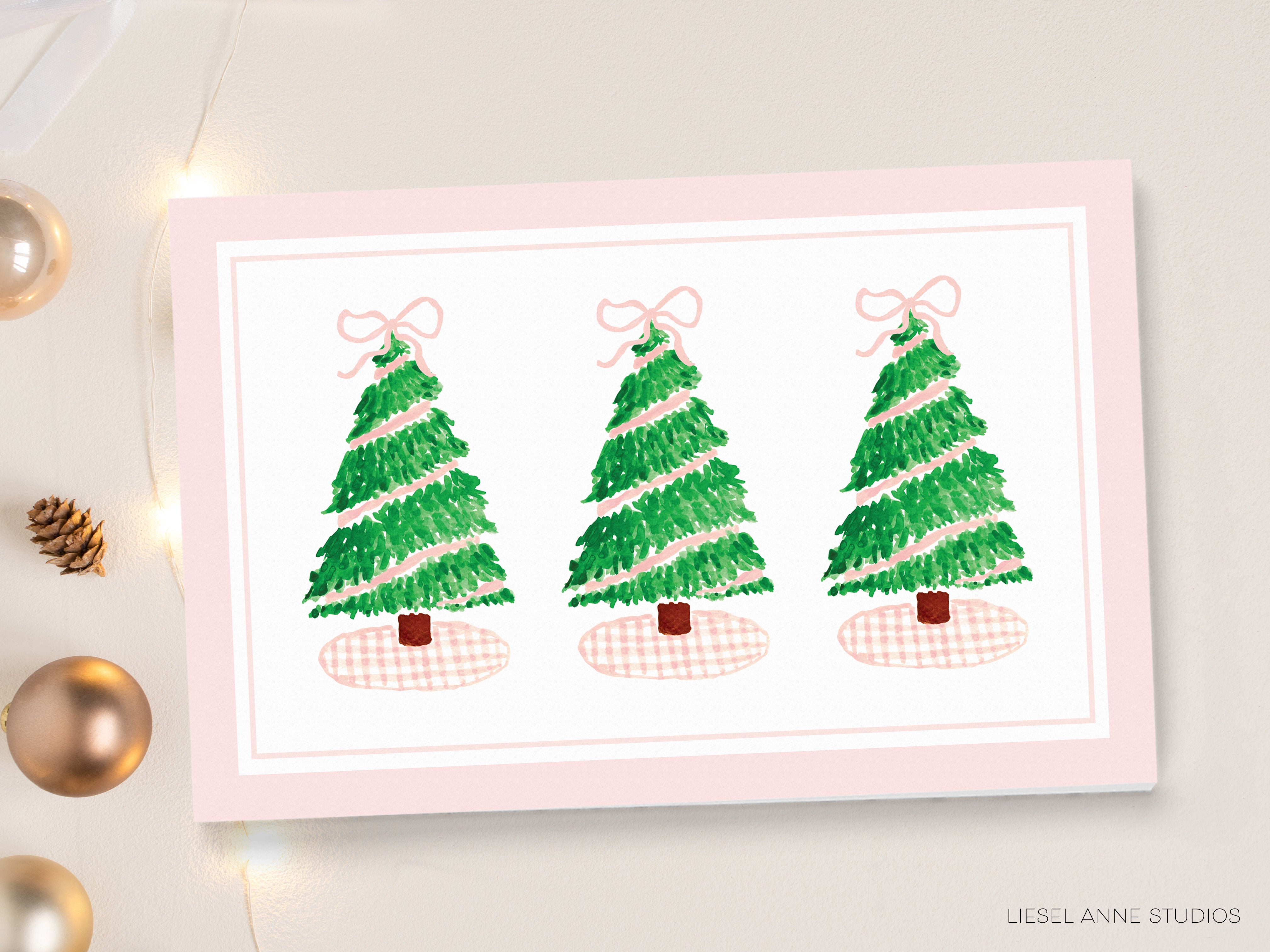 A pink greeting card with three Christmas trees and pink bows on top, placed on a white surface with Christmas ornaments and lights in the background.