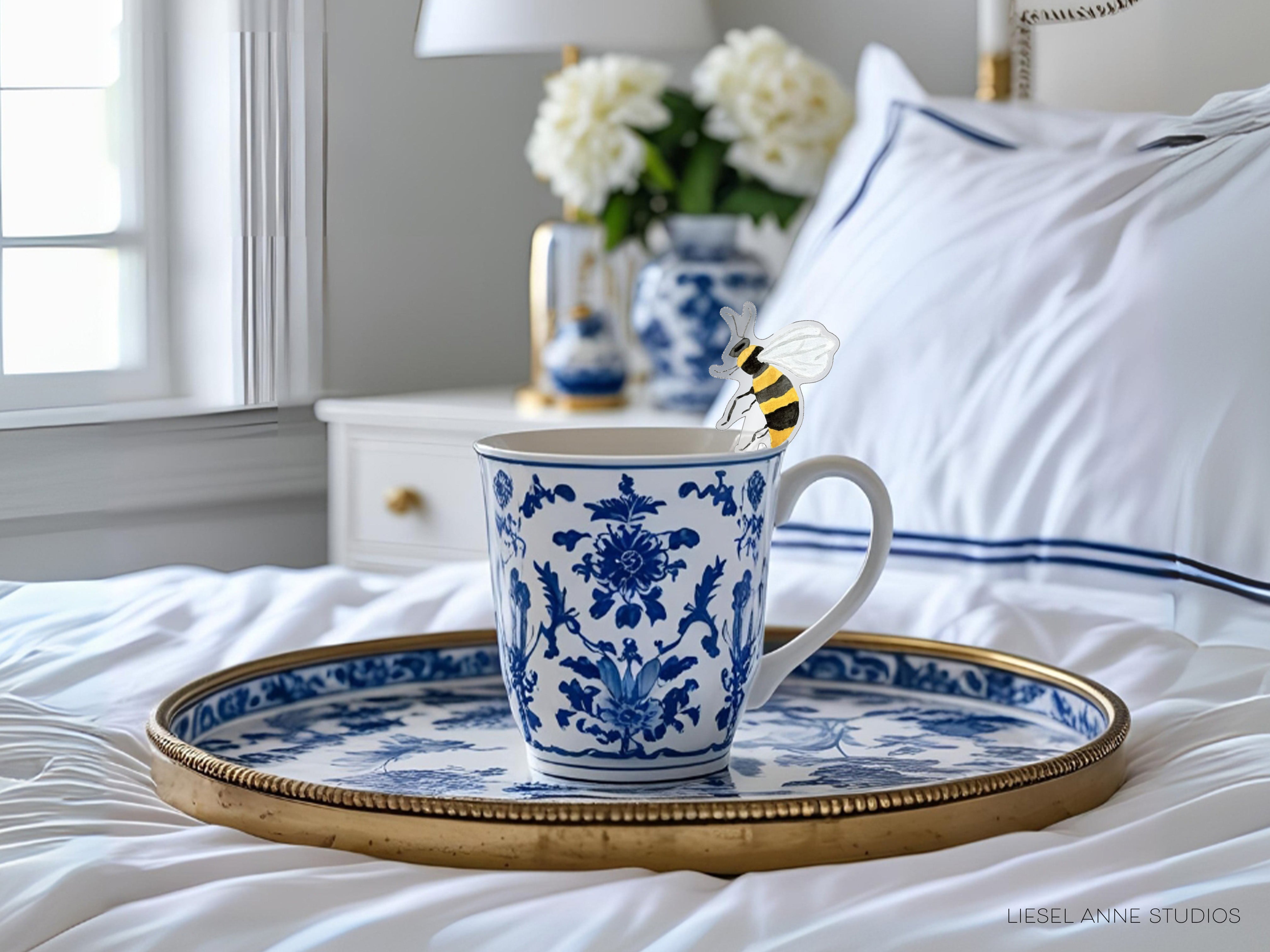 A blue and white patterned mug with a bee figurine in it sits on a tray, placed on a bed with white sheets and a white pillow.