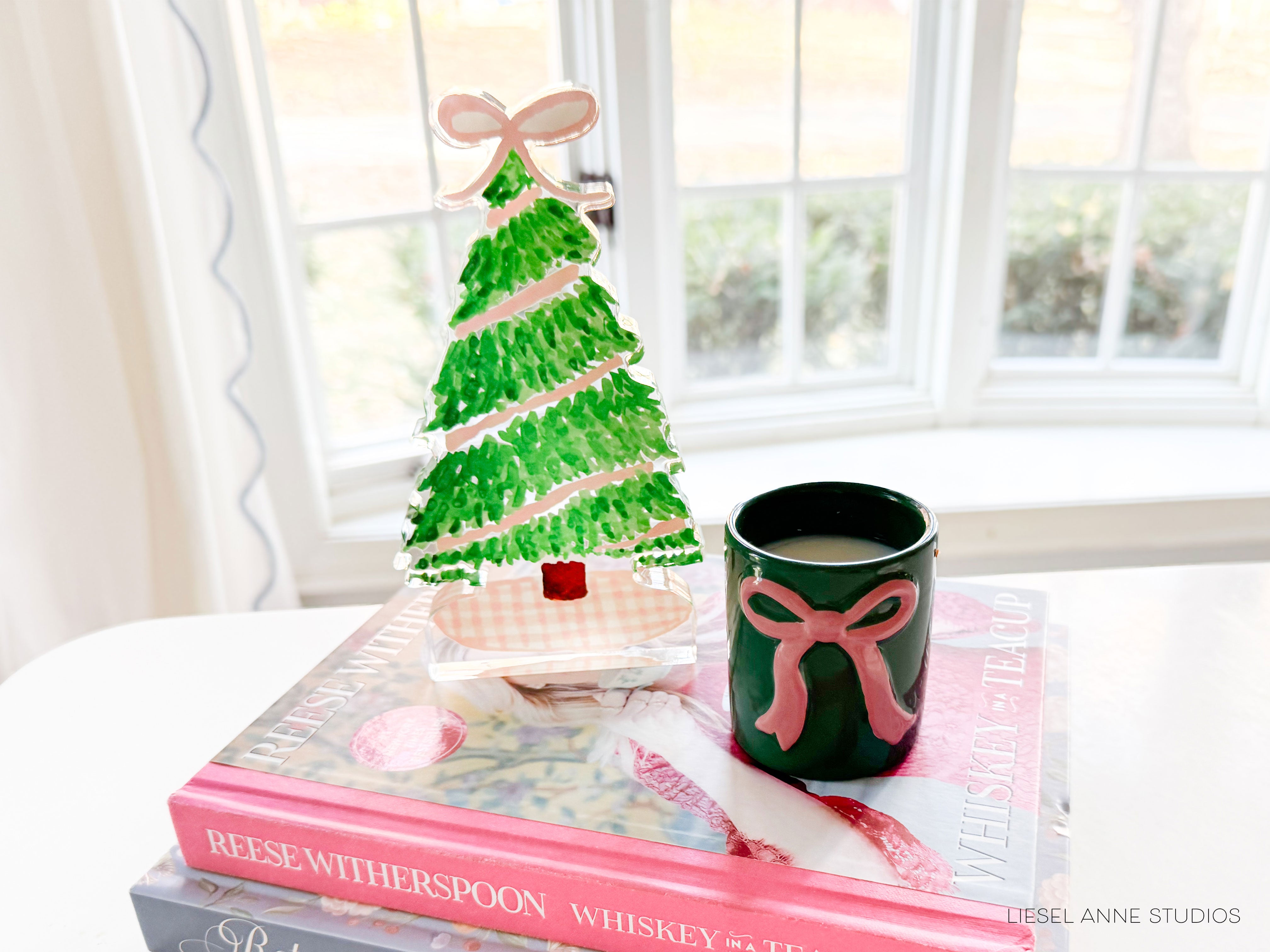 A green Christmas tree-shaped ornament with a pink bow sits on top of a stack of books, accompanied by a green mug with a pink bow.
