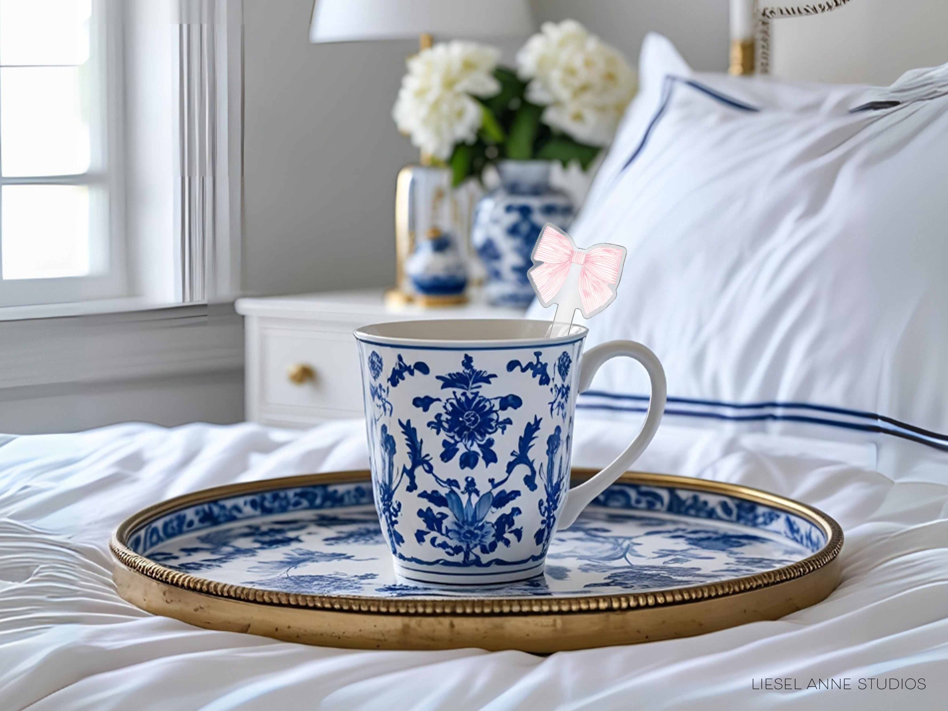 A blue and white patterned mug with a pink bow on a tray, placed on a bed with white sheets and a white pillow.