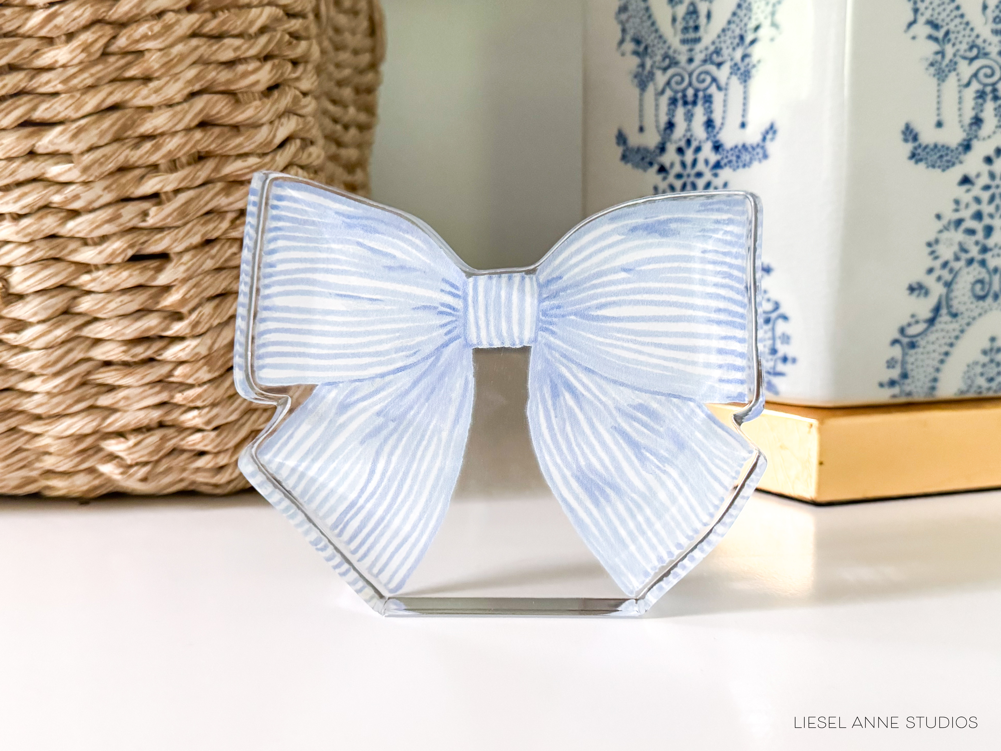 A decorative bow made of blue and white striped material is displayed on a white surface, with a woven basket and a blue and white patterned vase in the background.