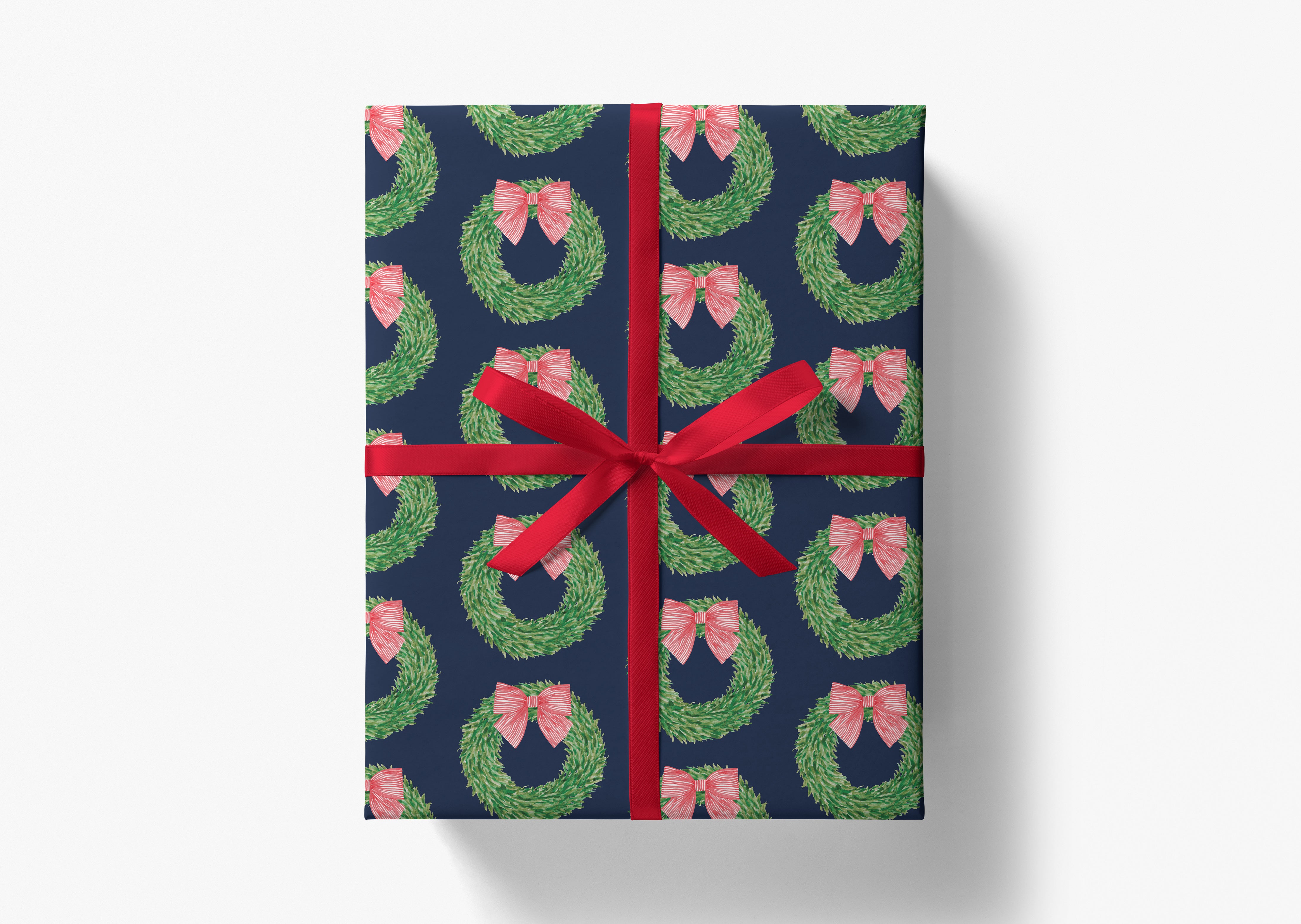 A gift box wrapped in a navy blue wrapping paper with a red ribbon and a repeating pattern of green wreaths with pink bows.
