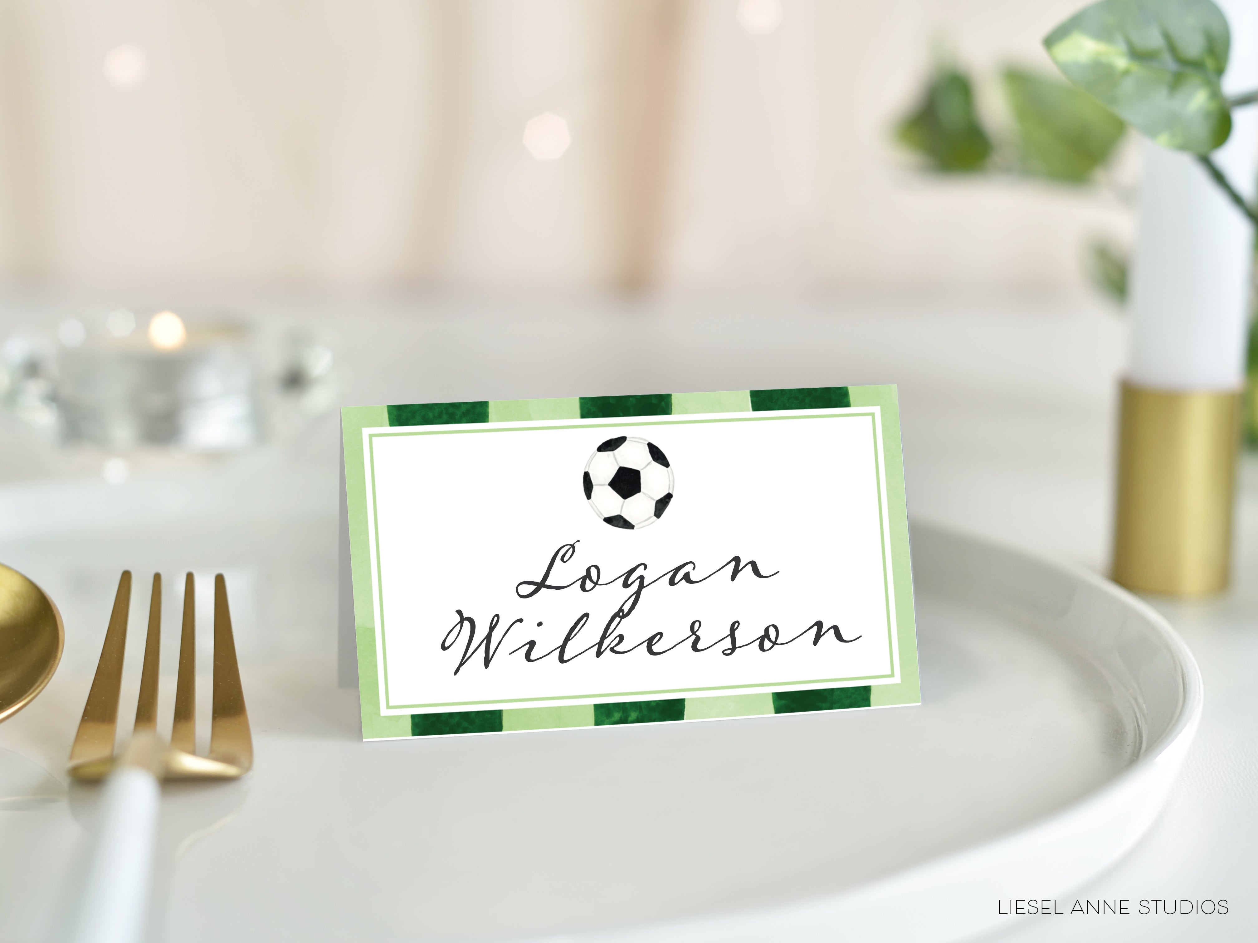 A green and white name card with a soccer ball design, placed on a white plate with a gold fork and knife.
