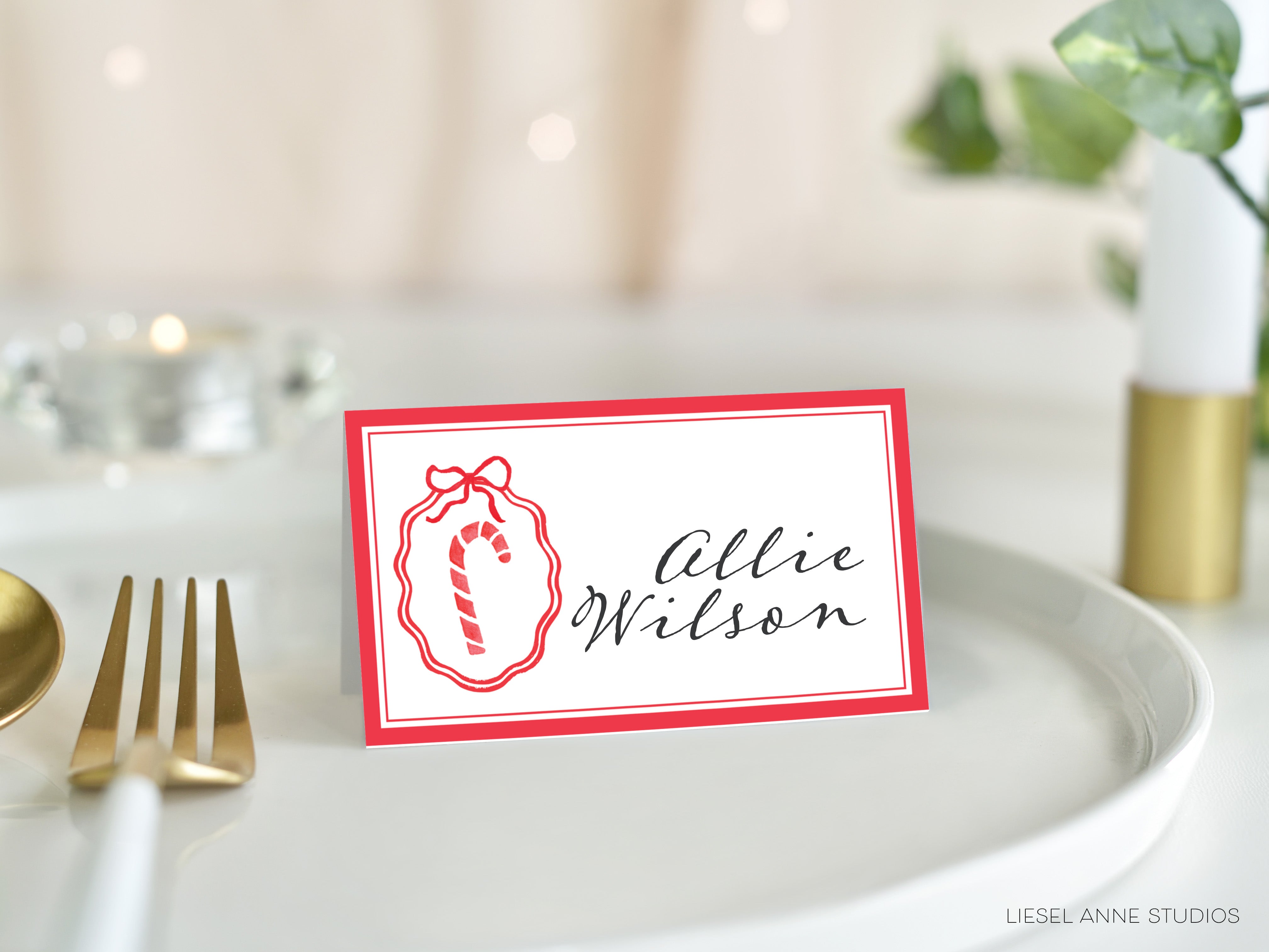 A red and white name card with a red ribbon and a candy cane design, placed on a white plate with a gold fork and spoon.