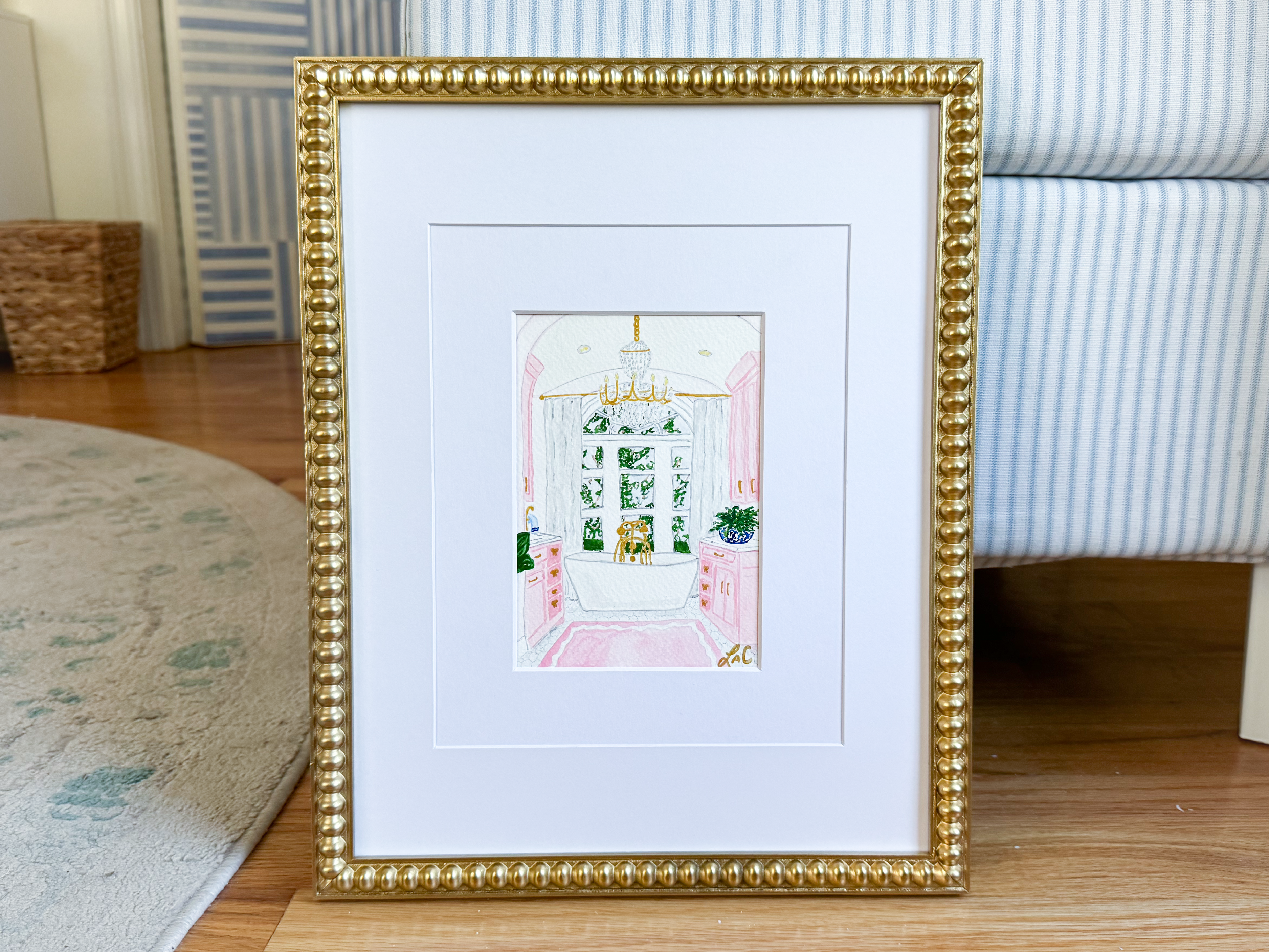 A framed painting depicting a bathroom interior with a pink and white color scheme and a chandelier is displayed on a wooden floor.