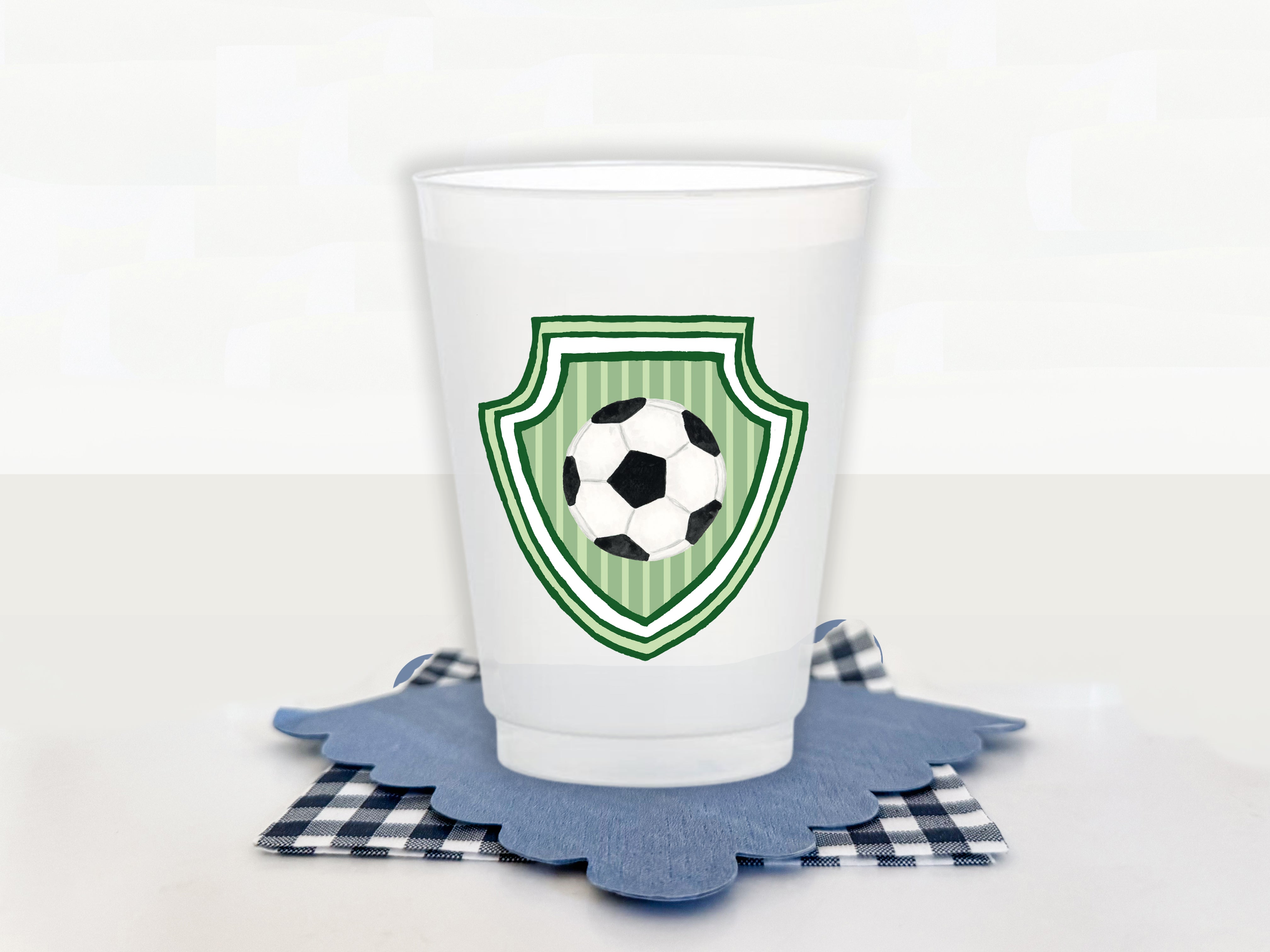 A white plastic cup with a soccer ball design on it, placed on a blue coaster with a checkered pattern.