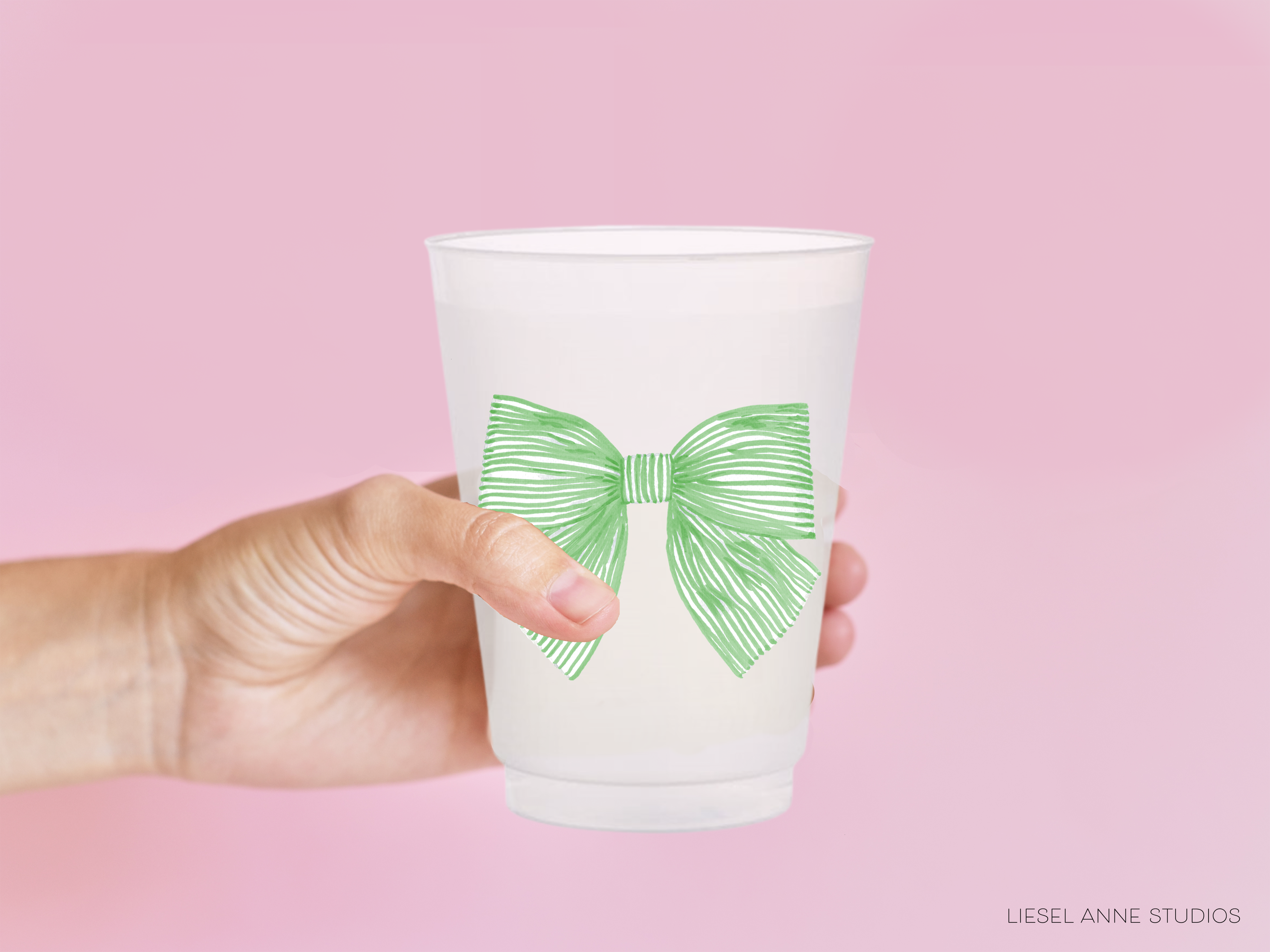 A hand holding a white plastic cup with a green bow on it against a pink background.