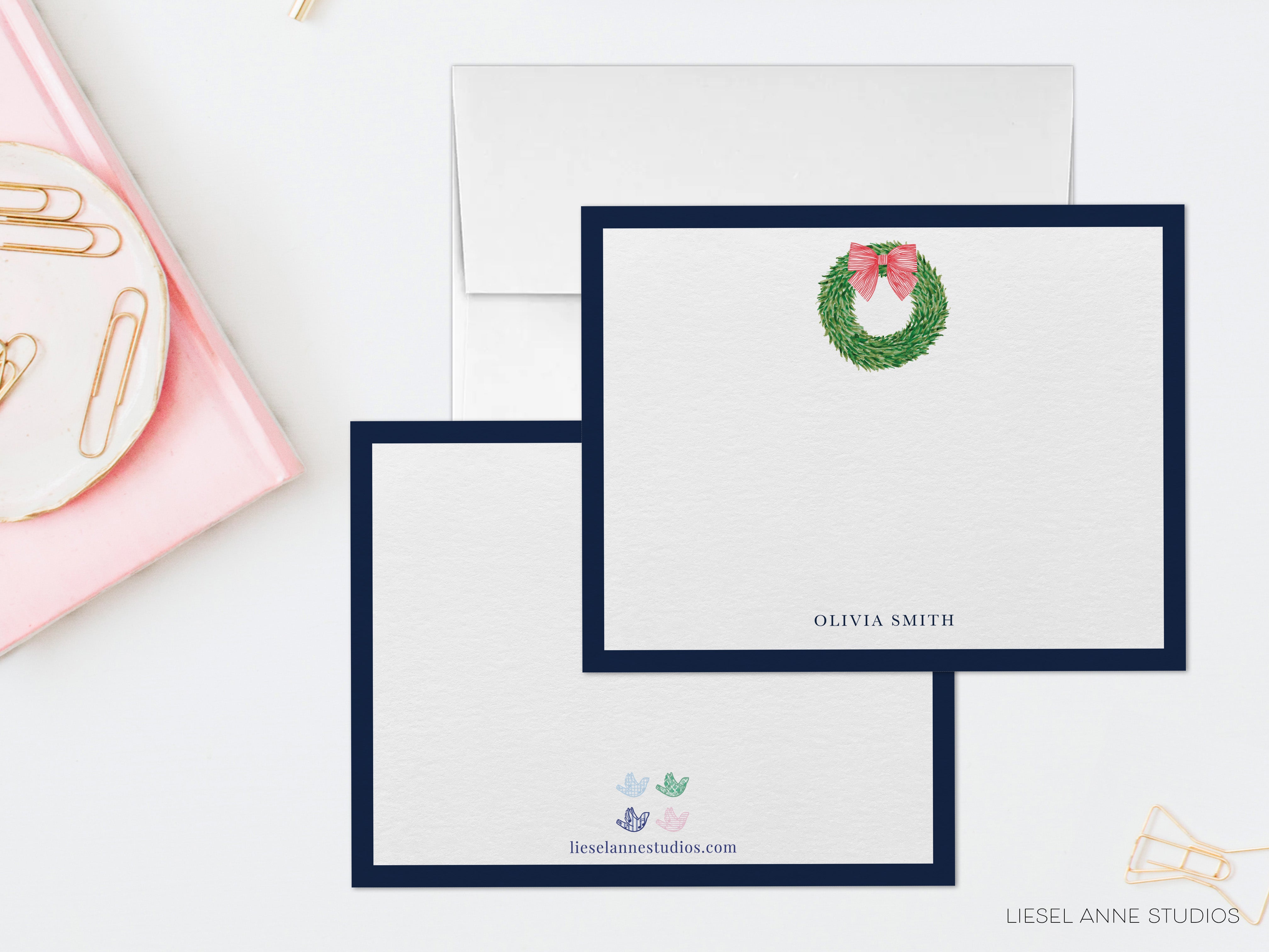 two blank greeting cards with a wreath design, placed on a white surface alongside a pink plate with gold clips.