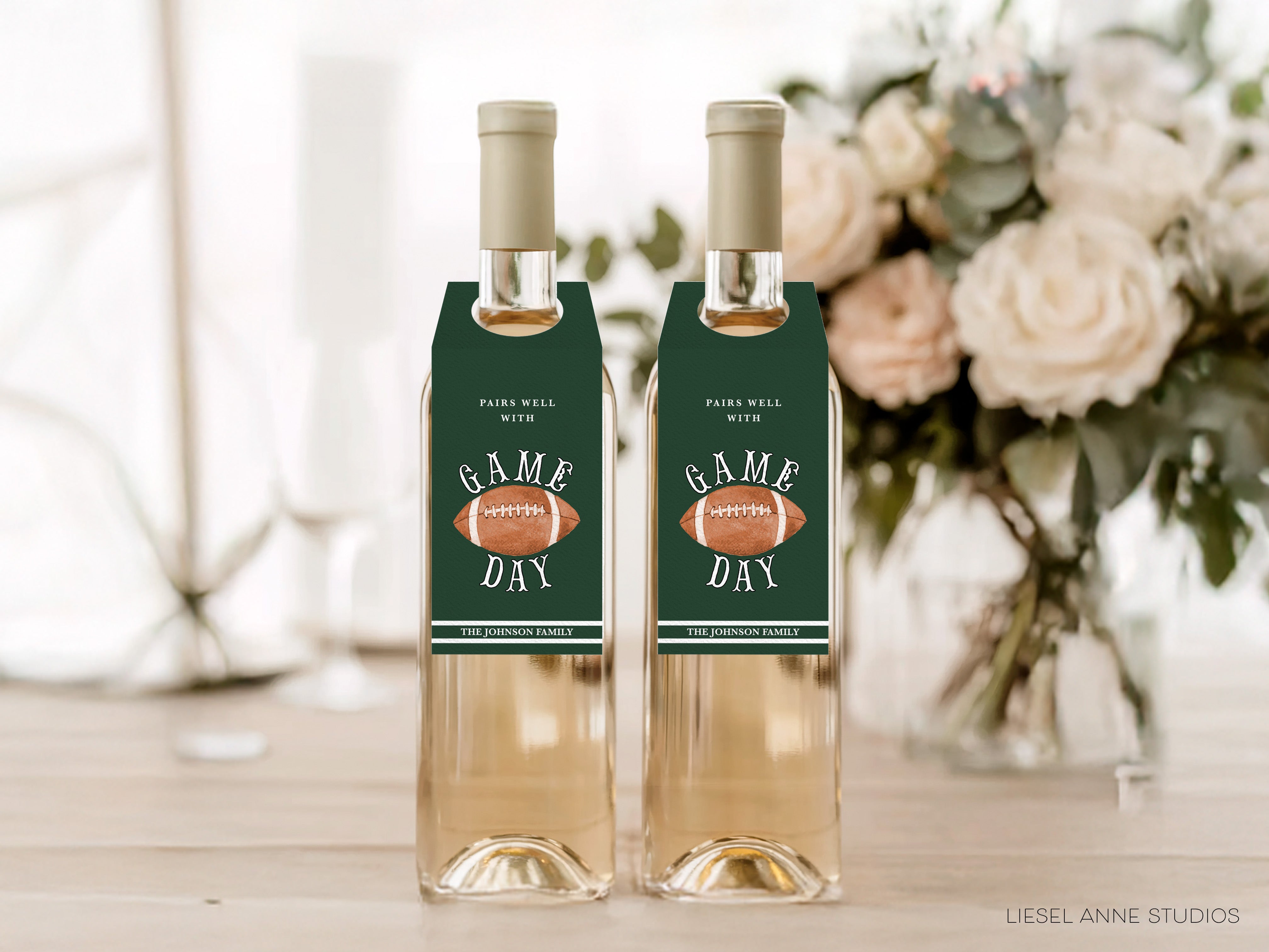 Two bottles of wine with green labels that say "GAME DAY" and "PAIRS WELL WITH", placed on a wooden surface in front of a vase of white flowers.