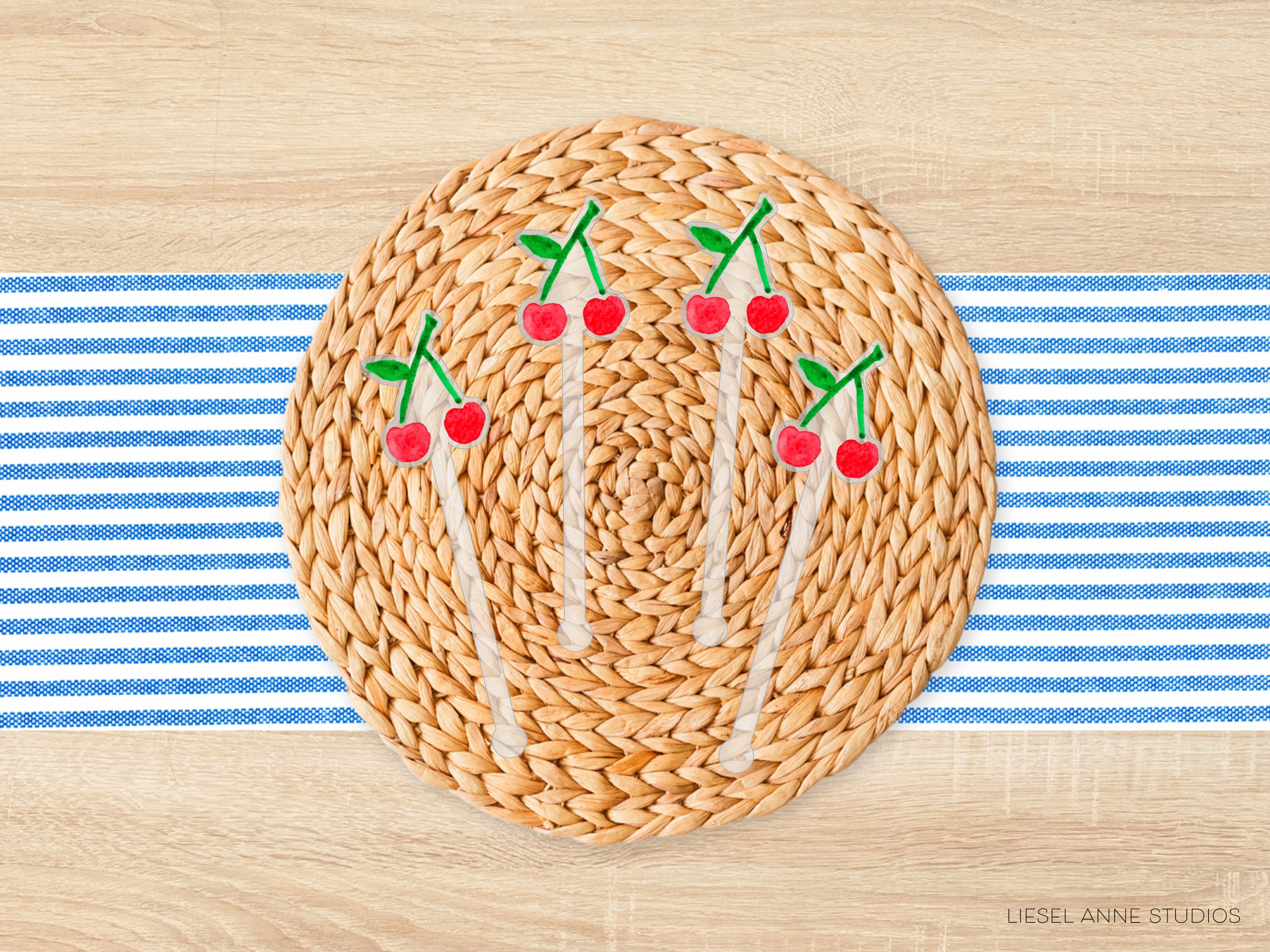 A woven placemat with a circular arrangement of red and green cherries on top, placed on a wooden surface with blue and white striped fabric.