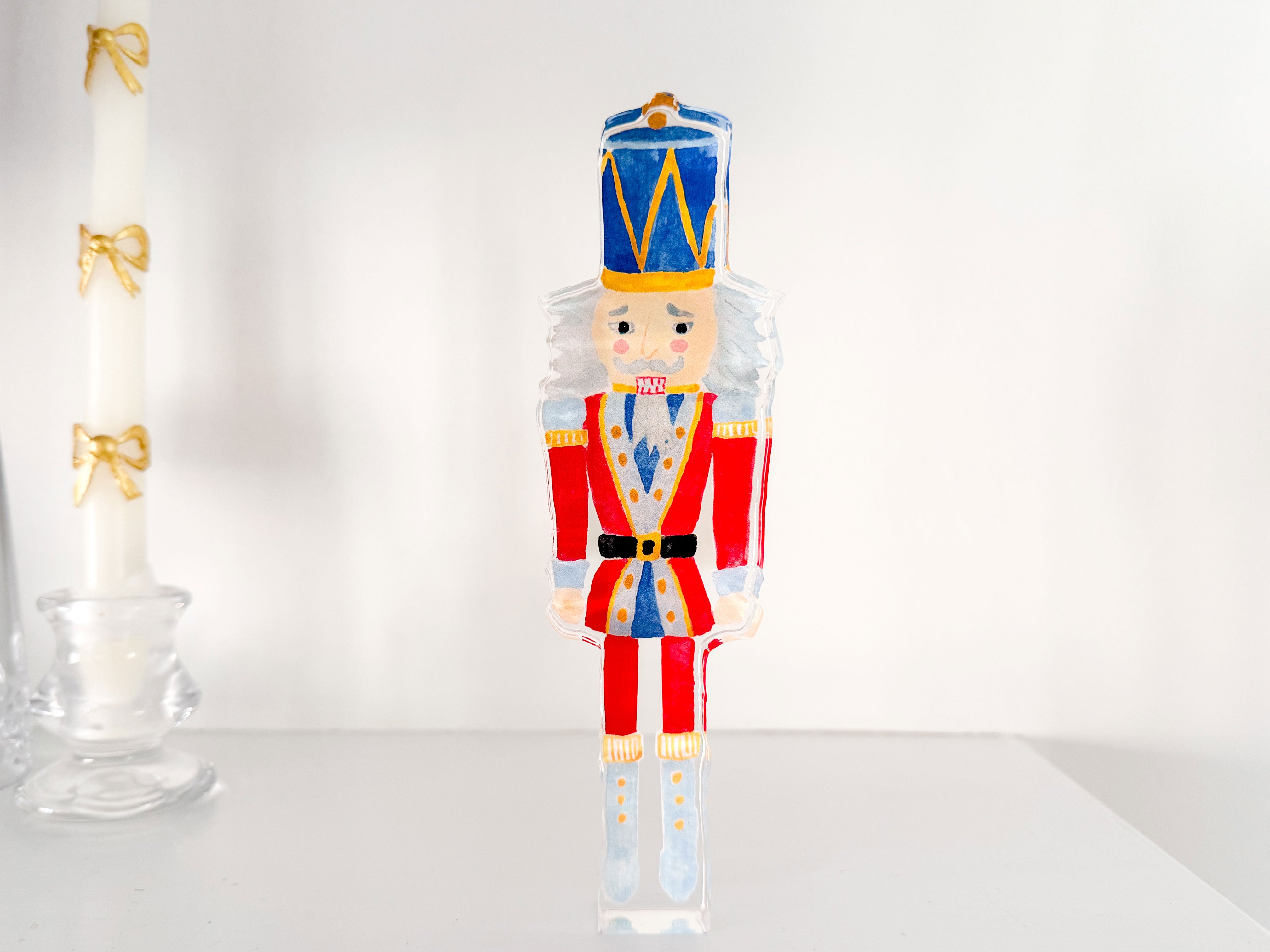 A colorful nutcracker figurine is displayed on a white surface, with a white candle and a glass candlestick holder visible in the background.
