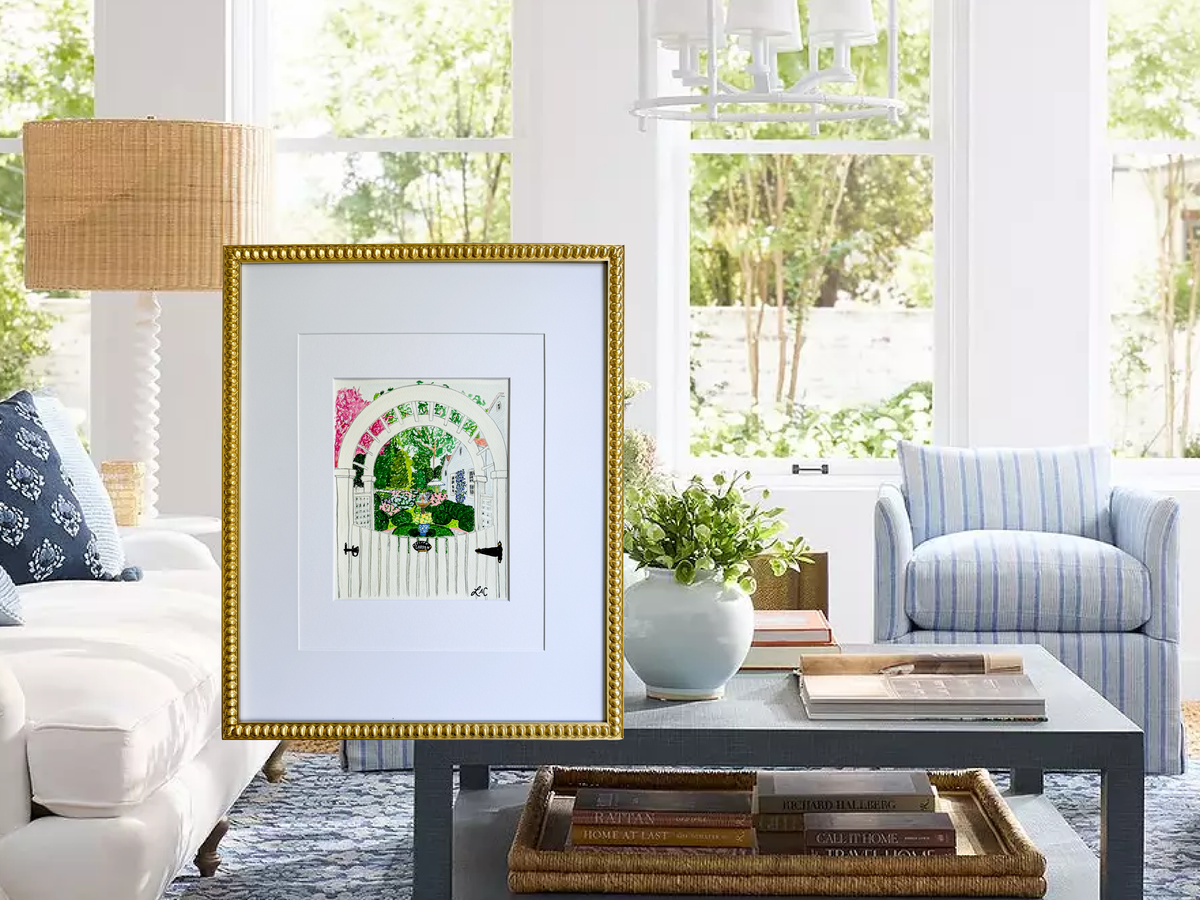 a cozy living room with a white sofa, a blue armchair, and a coffee table with books and a vase. A framed artwork of a garden gate is displayed on the coffee table, and a lamp is positioned on the left side of the room.