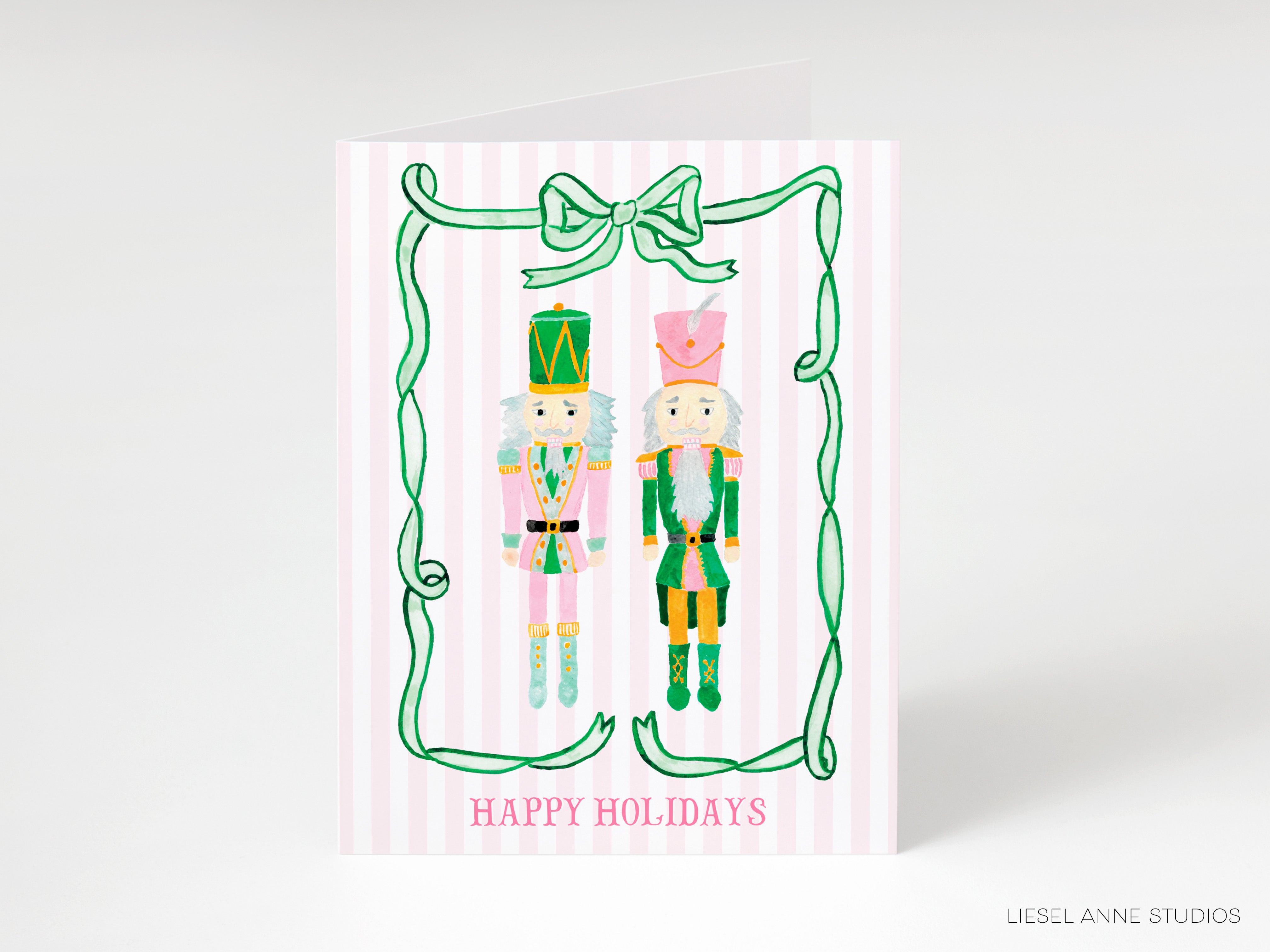 A greeting card featuring two nutcracker characters, one in a pink outfit and the other in a green outfit, standing together with a green ribbon bow. The card has the text "Happy Holidays" written on it.