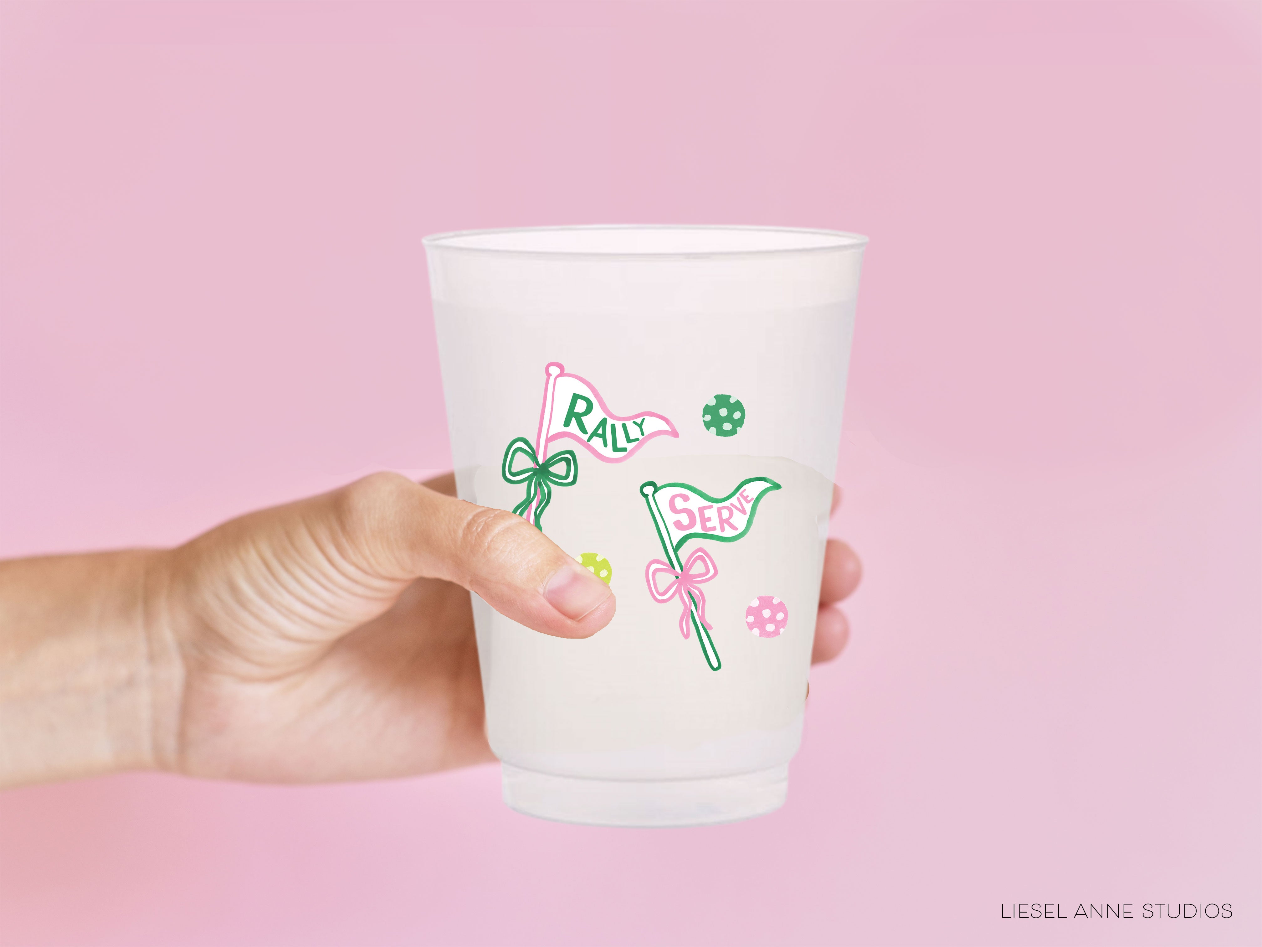 A hand holding a white plastic cup with a design featuring green and pink flags and flowers against a pink background.