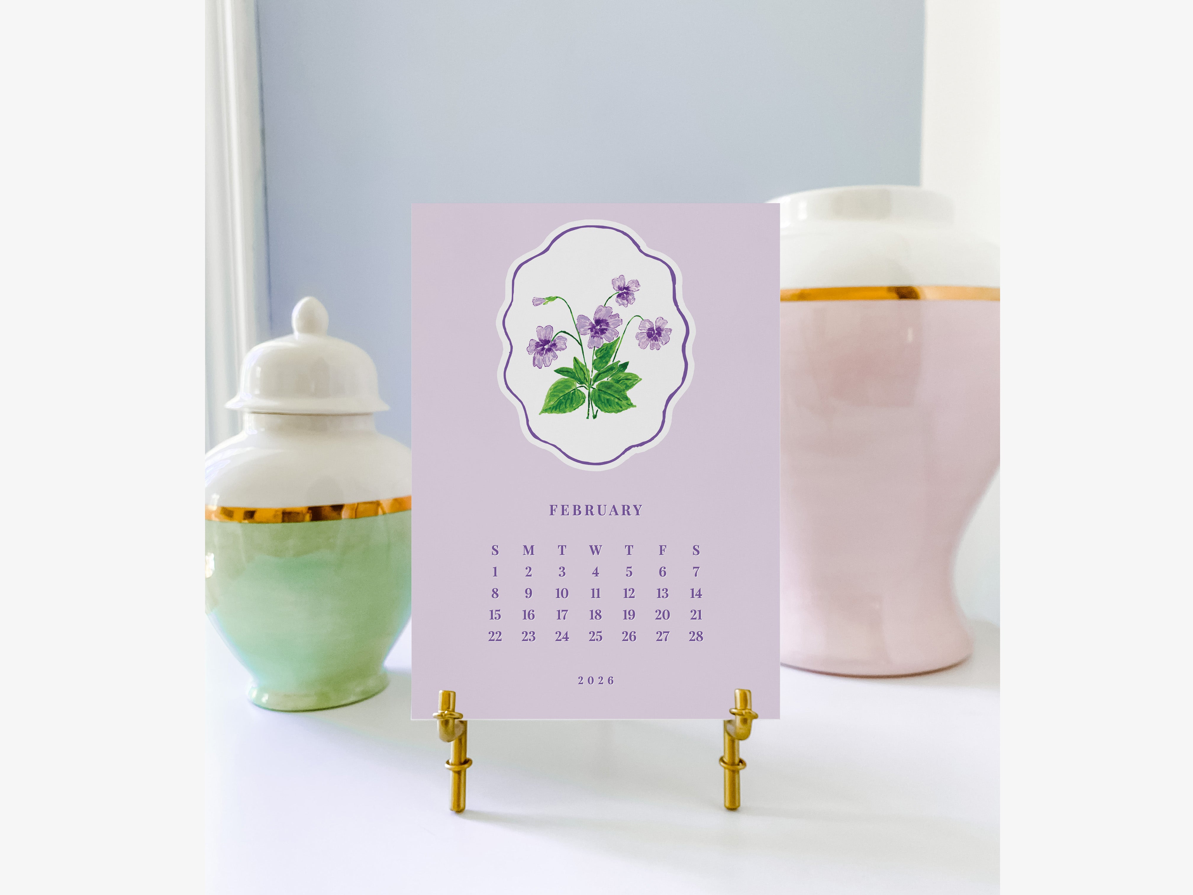 A calendar for February 2016 with a purple background and a white border, featuring a floral design. The calendar is placed on a stand between two ceramic vases, one pink and one green, against a light blue background.