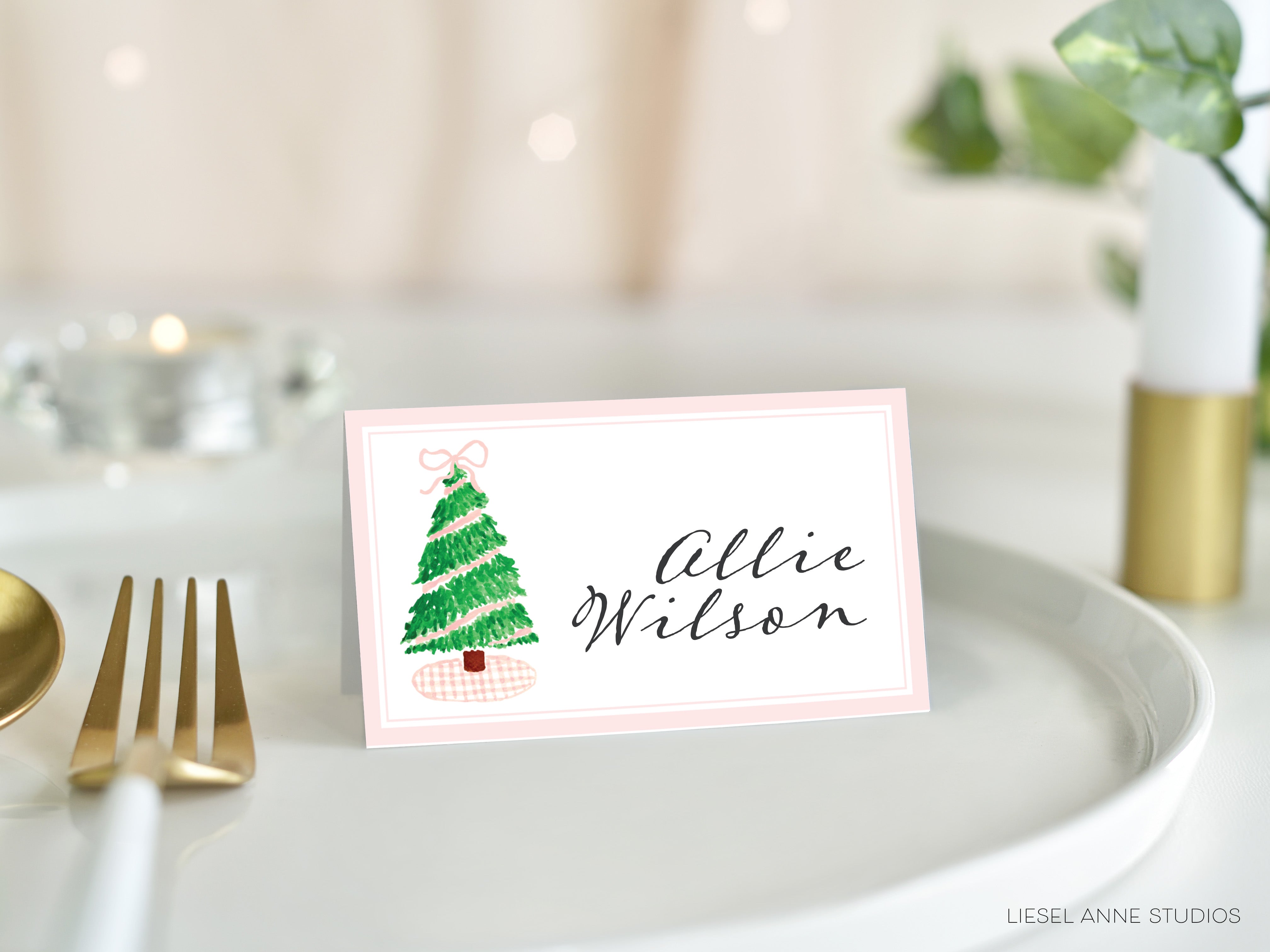 A pink name card with a Christmas tree illustration and the name "Allie Wilson" written on it, placed on a white plate.