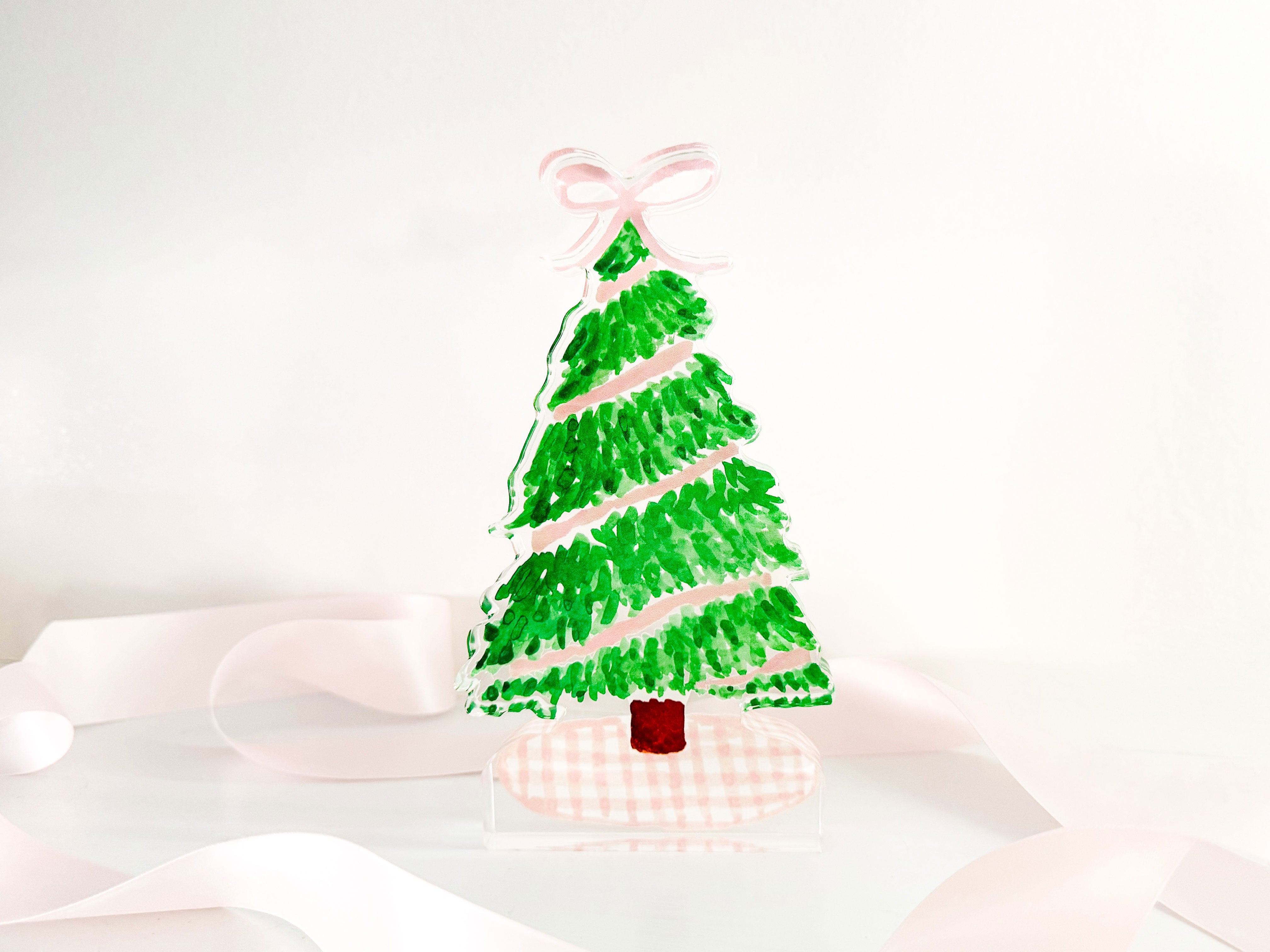 A hand-painted Christmas tree with green and pink colors stands on a pink ribbon, creating a festive and decorative scene.