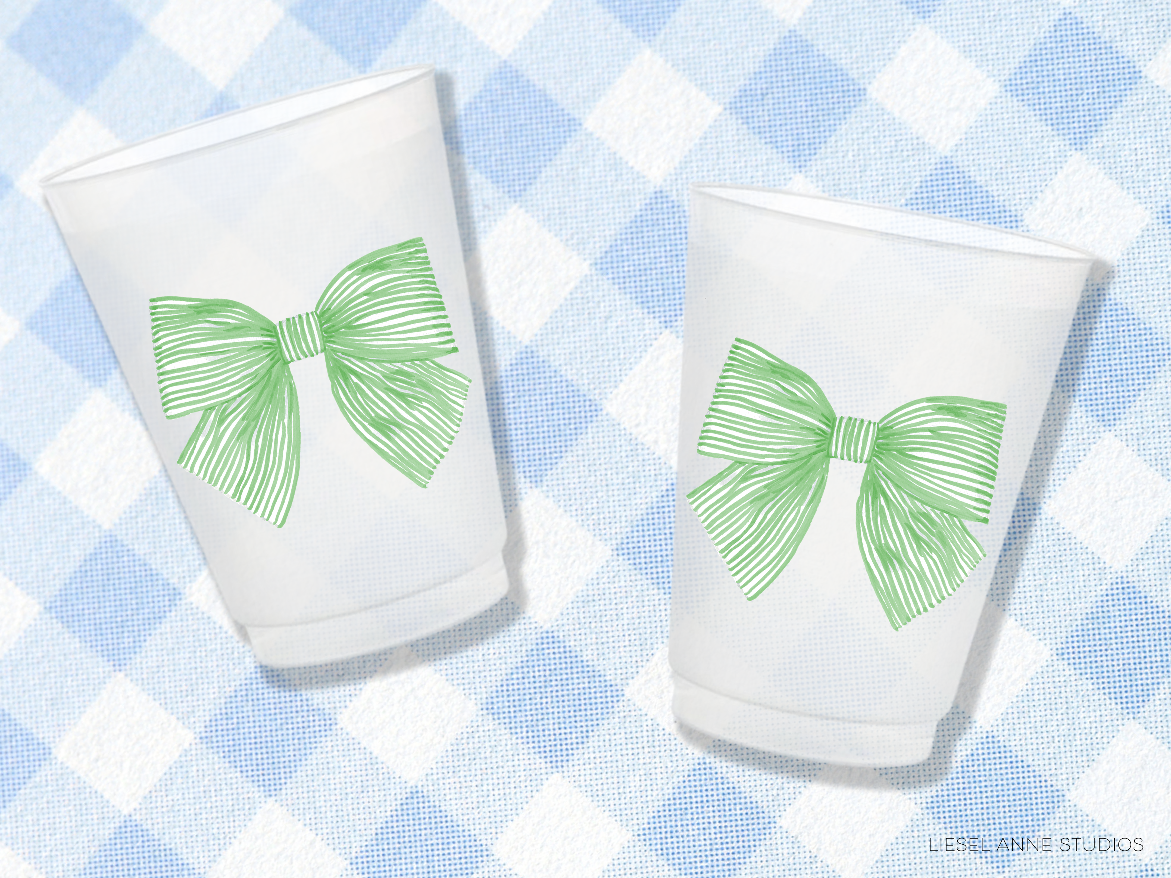 Two white cups with green bows on them, placed on a blue and white checkered tablecloth.
