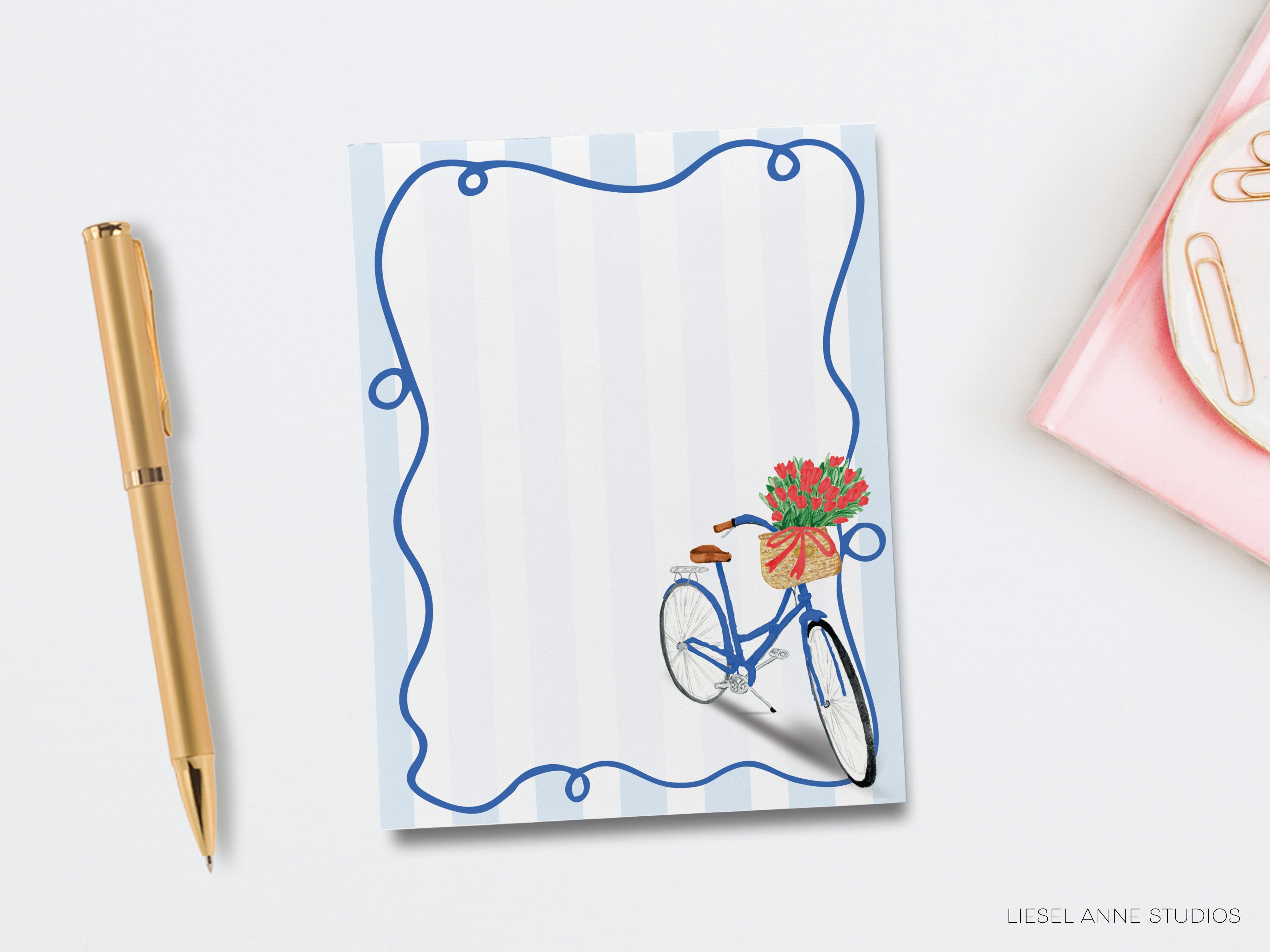 a blue bicycle with a basket of flowers, placed on a white background. There is also a gold pen and a pink object, possibly a notebook or a pen holder, located next to the bicycle.