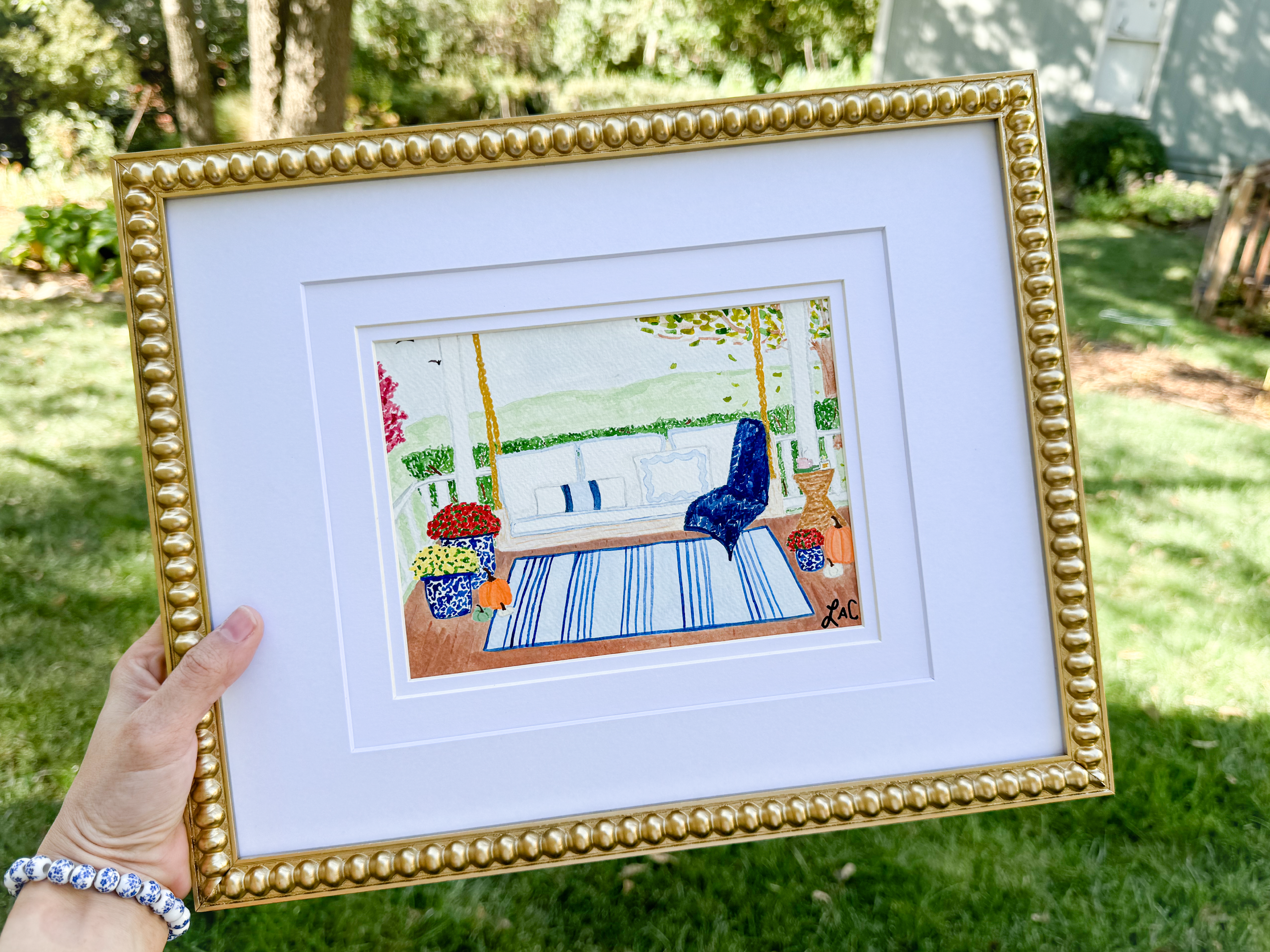 A hand holding a framed painting of a porch with a blue chair, potted plants, and a rug.
