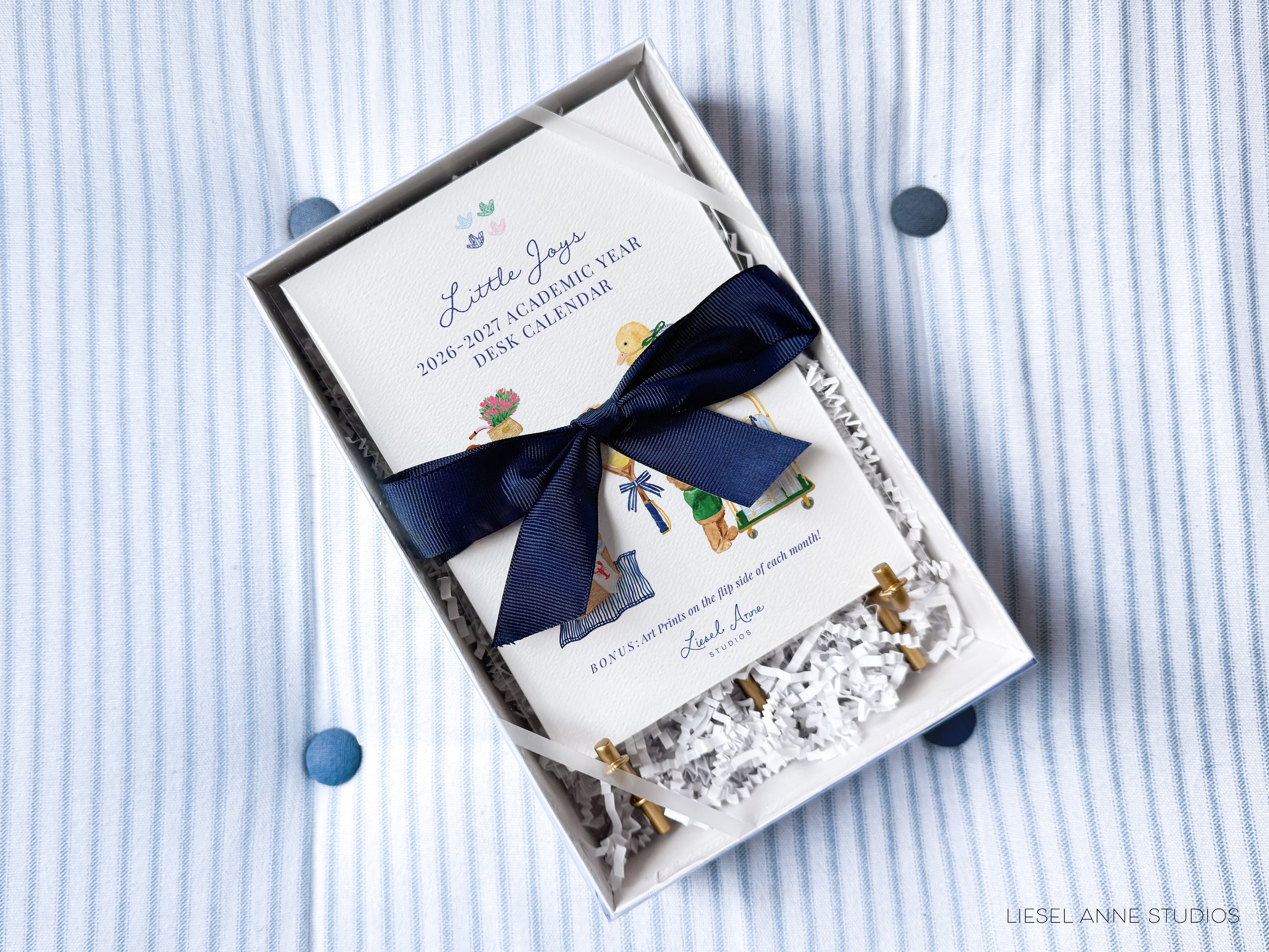A white box containing a white card with illustrations and a blue ribbon tied around it, placed on a striped fabric background.