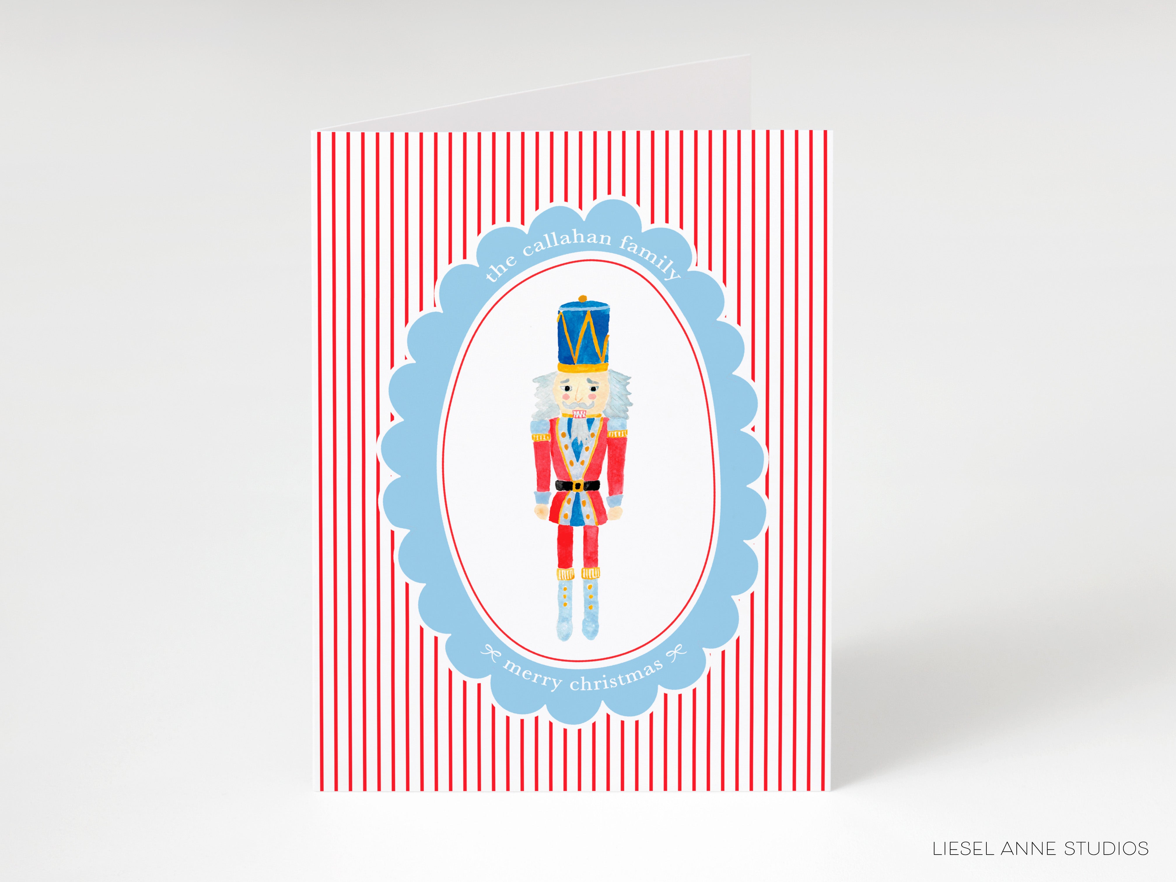 A Christmas card featuring a nutcracker figure in a blue and red outfit, standing in the center of a blue and red striped background with a scalloped edge.