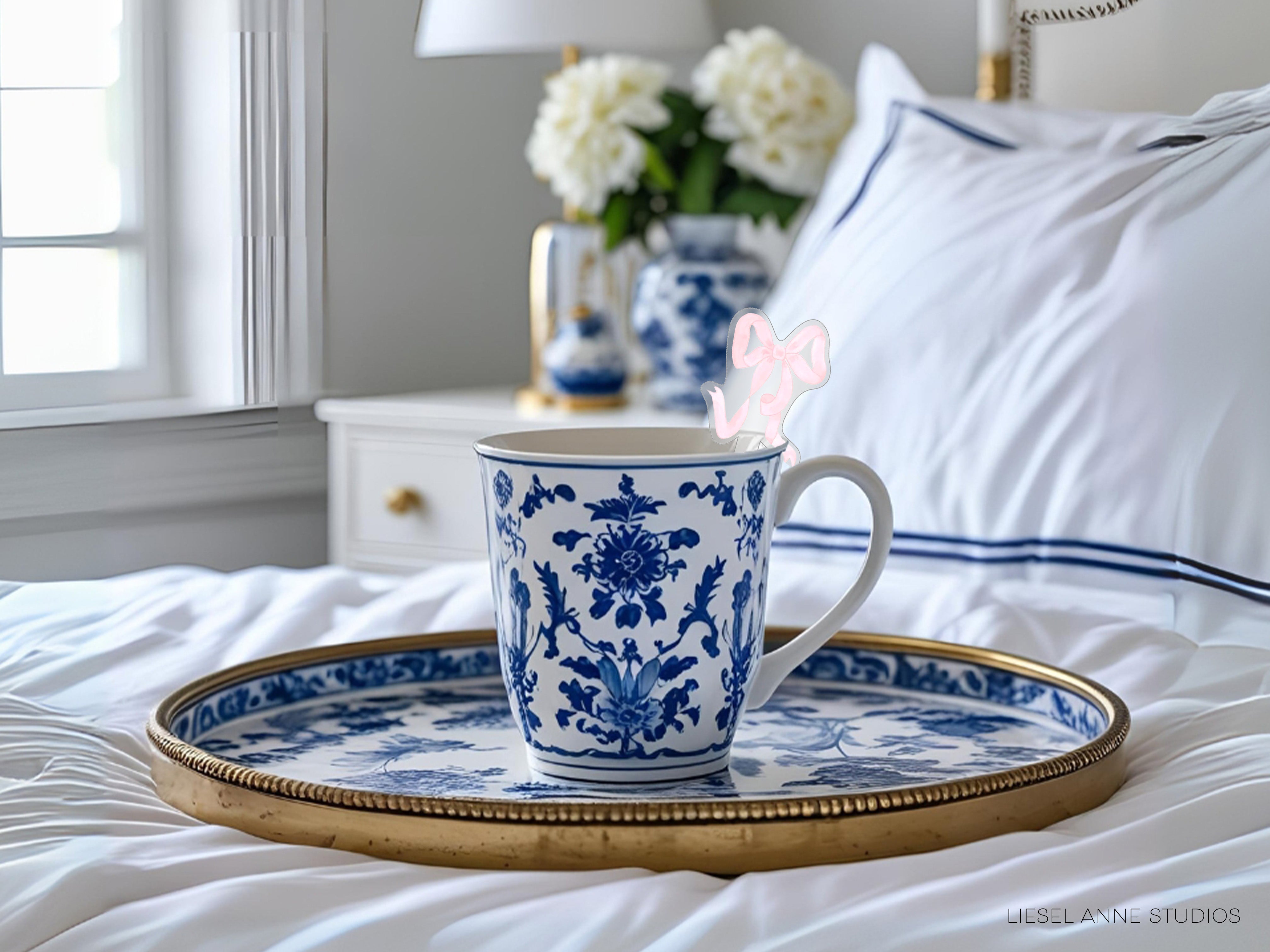 A blue and white patterned coffee mug sits on a gold tray, with a vase of white flowers and a pink bow in the background.
