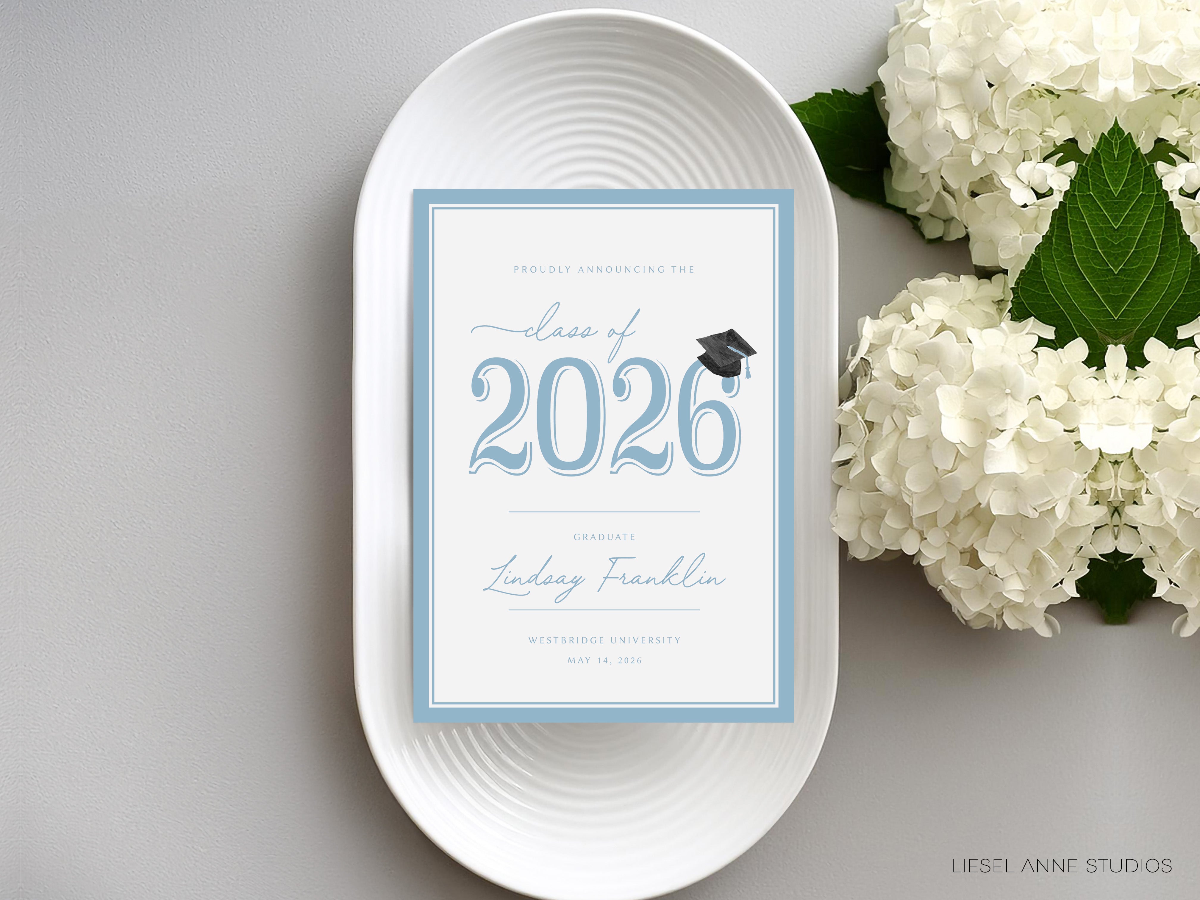 a graduation invitation card with the text "Class of 2026 Linday Franklin Westbridge University May 4, 2026" displayed on it, placed on a white plate next to a bouquet of white hydrangeas.