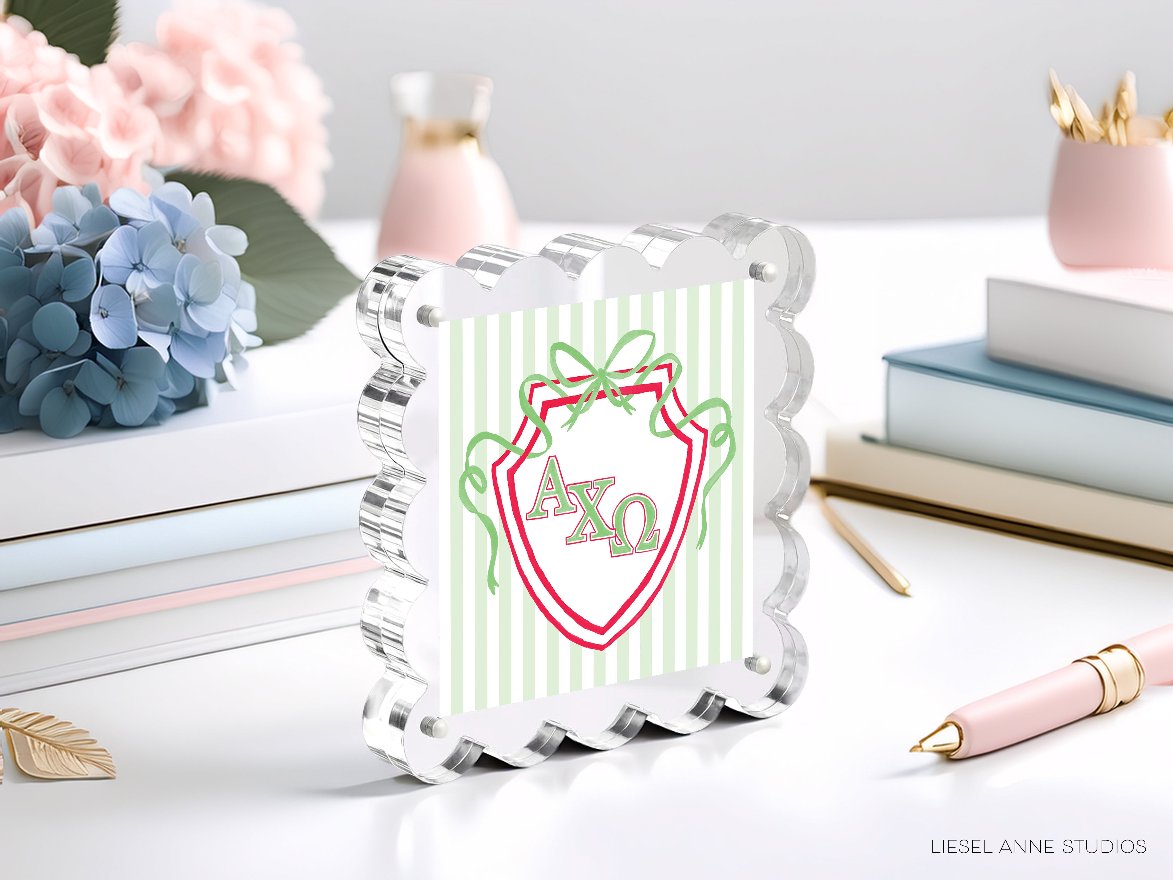 a clear glass frame with a decorative border, displaying a shield-shaped logo with the letters "AXQ" in green and red colors. The frame is placed on a table surrounded by various items, including a stack of books, a pink vase, and a pink pen.