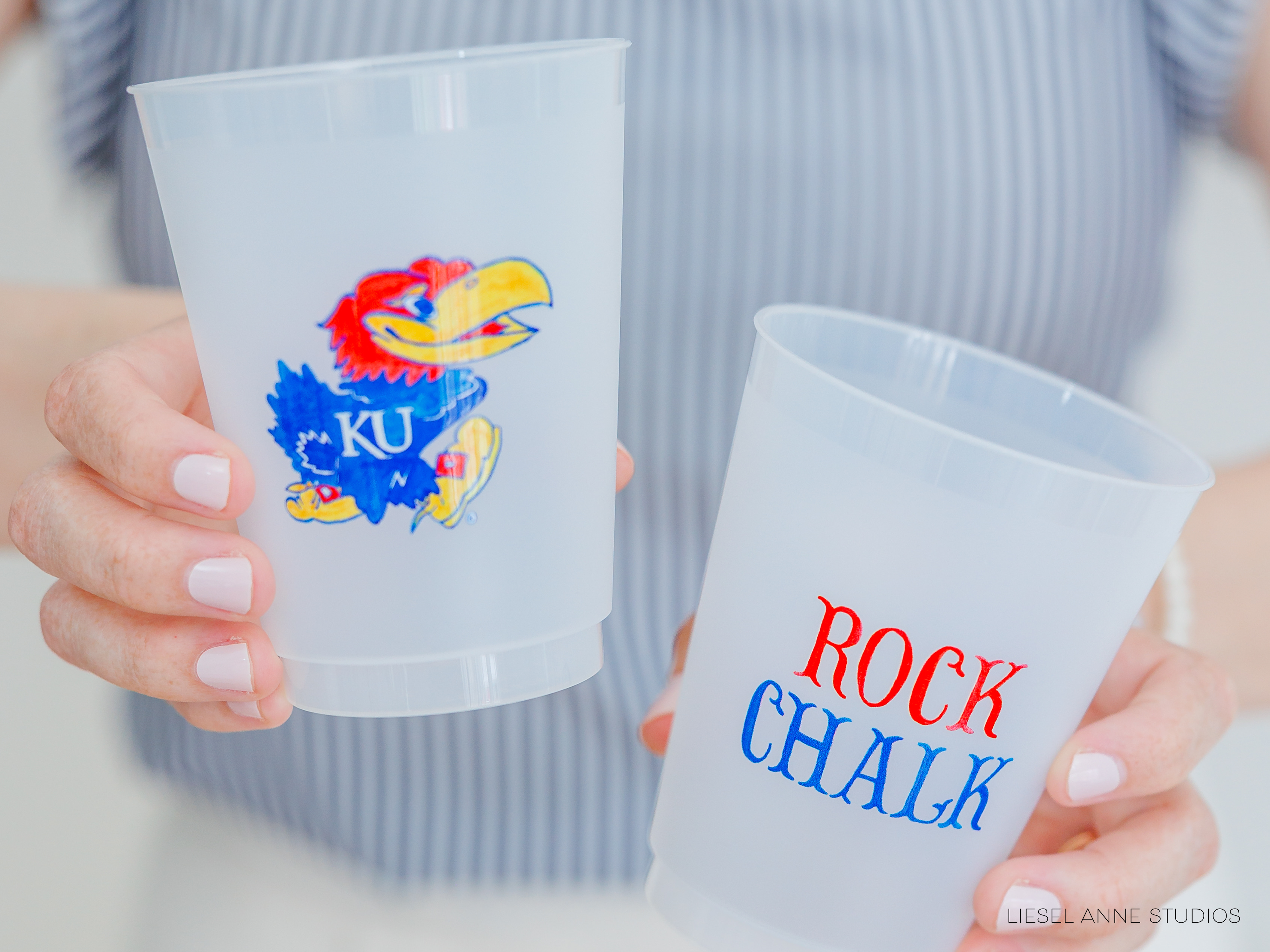 A person is holding two plastic cups, one with a colorful bird design and the other with the text "ROCK CHALK" printed on it.