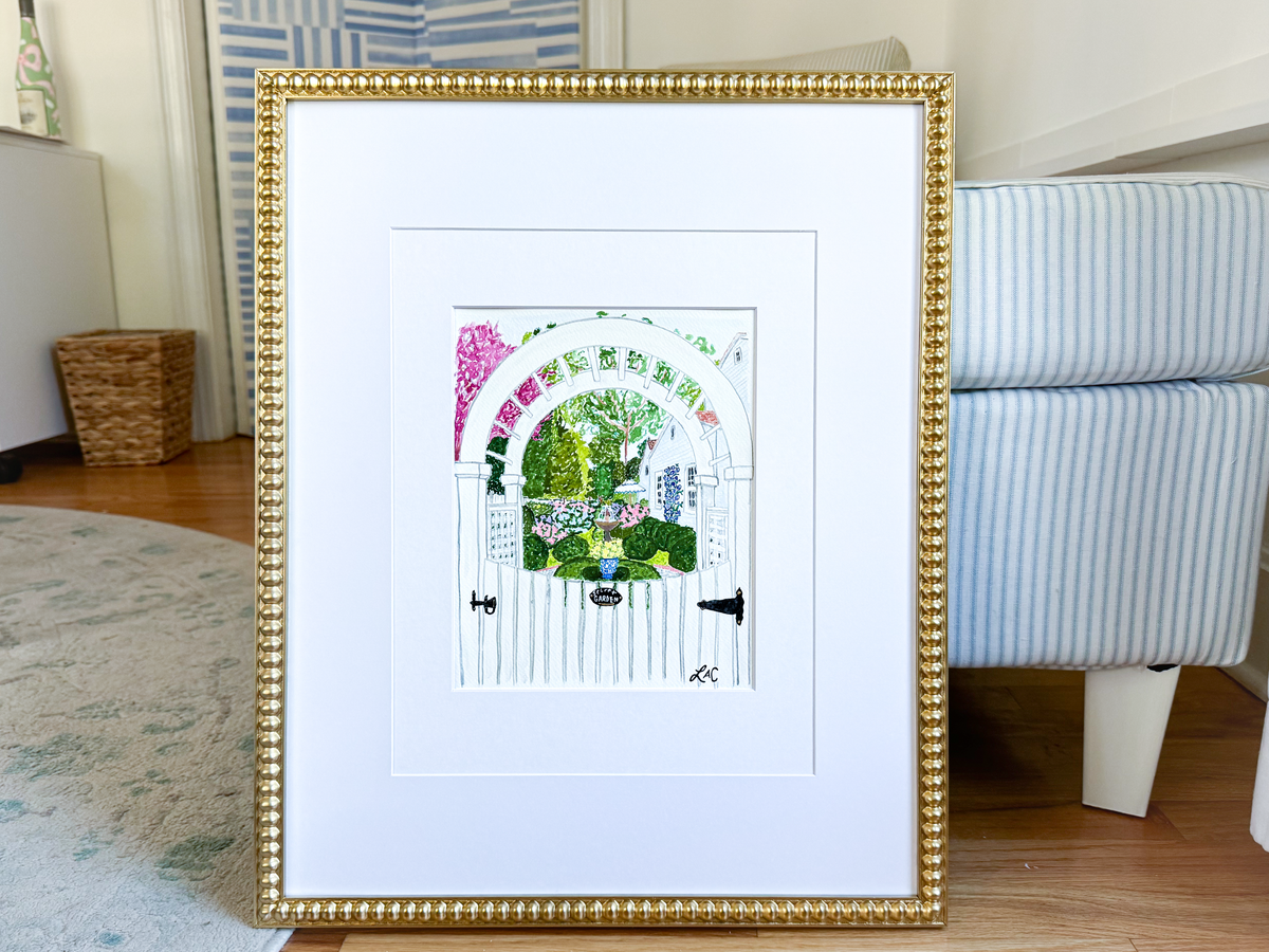 A framed print of a white wooden gate with a colorful garden scene is displayed on a wooden floor in a room with a blue and white striped chair.