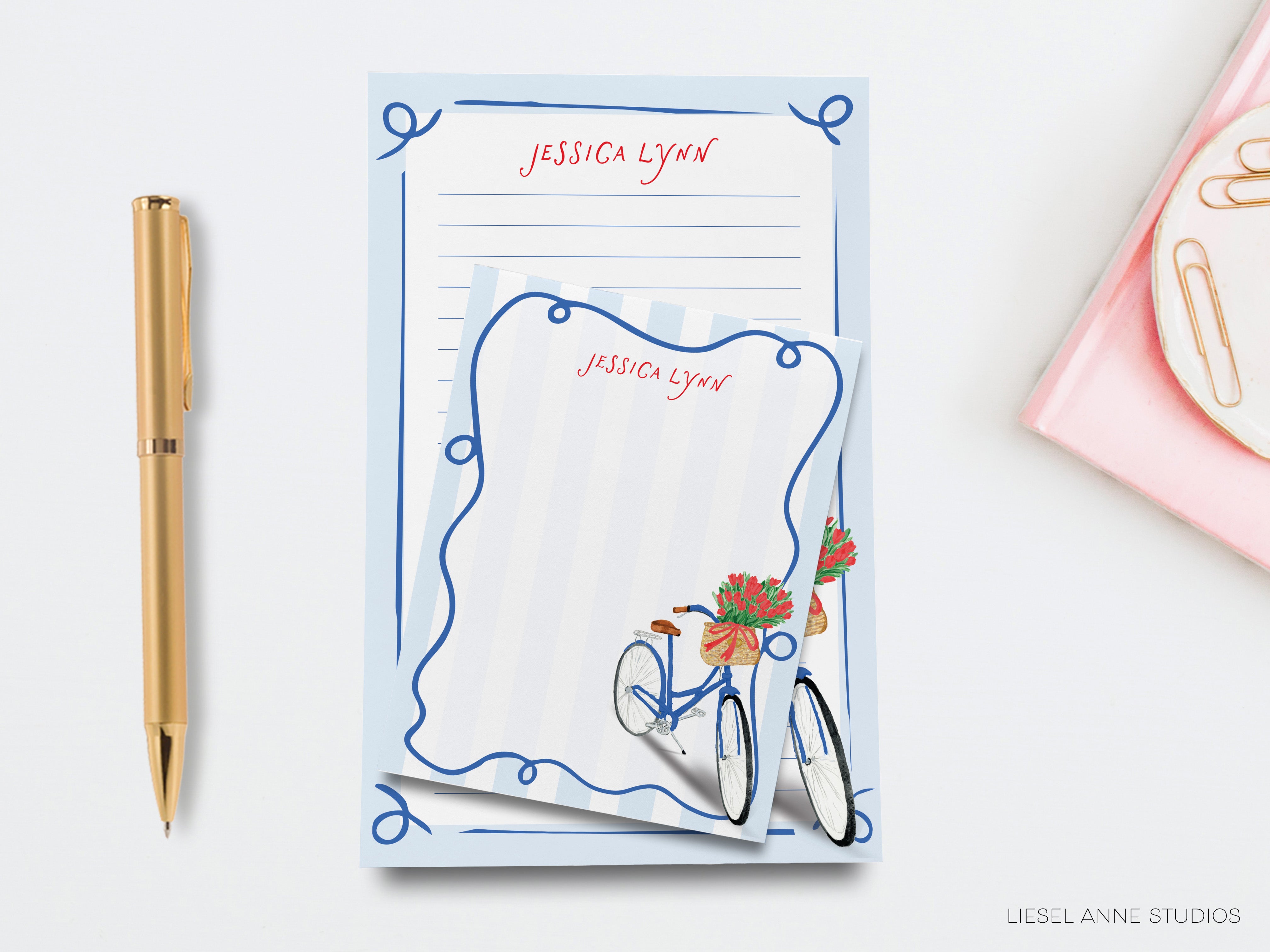 a blue notepad with a bicycle illustration, a gold pen, and a pink object on a white background.