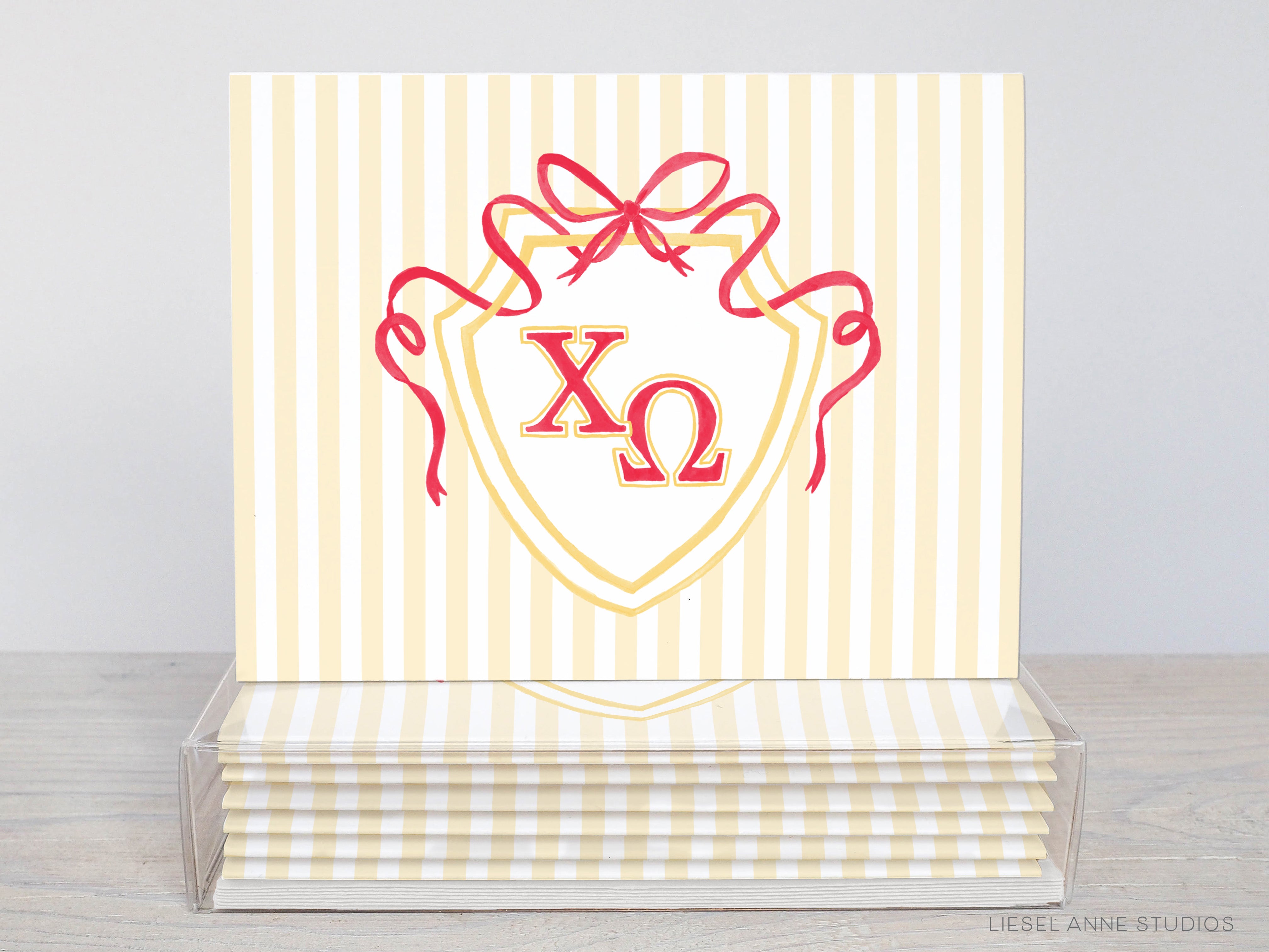 A stack of yellow and white striped greeting cards with a red ribbon and a shield-shaped design featuring the Greek letters omega and omega.