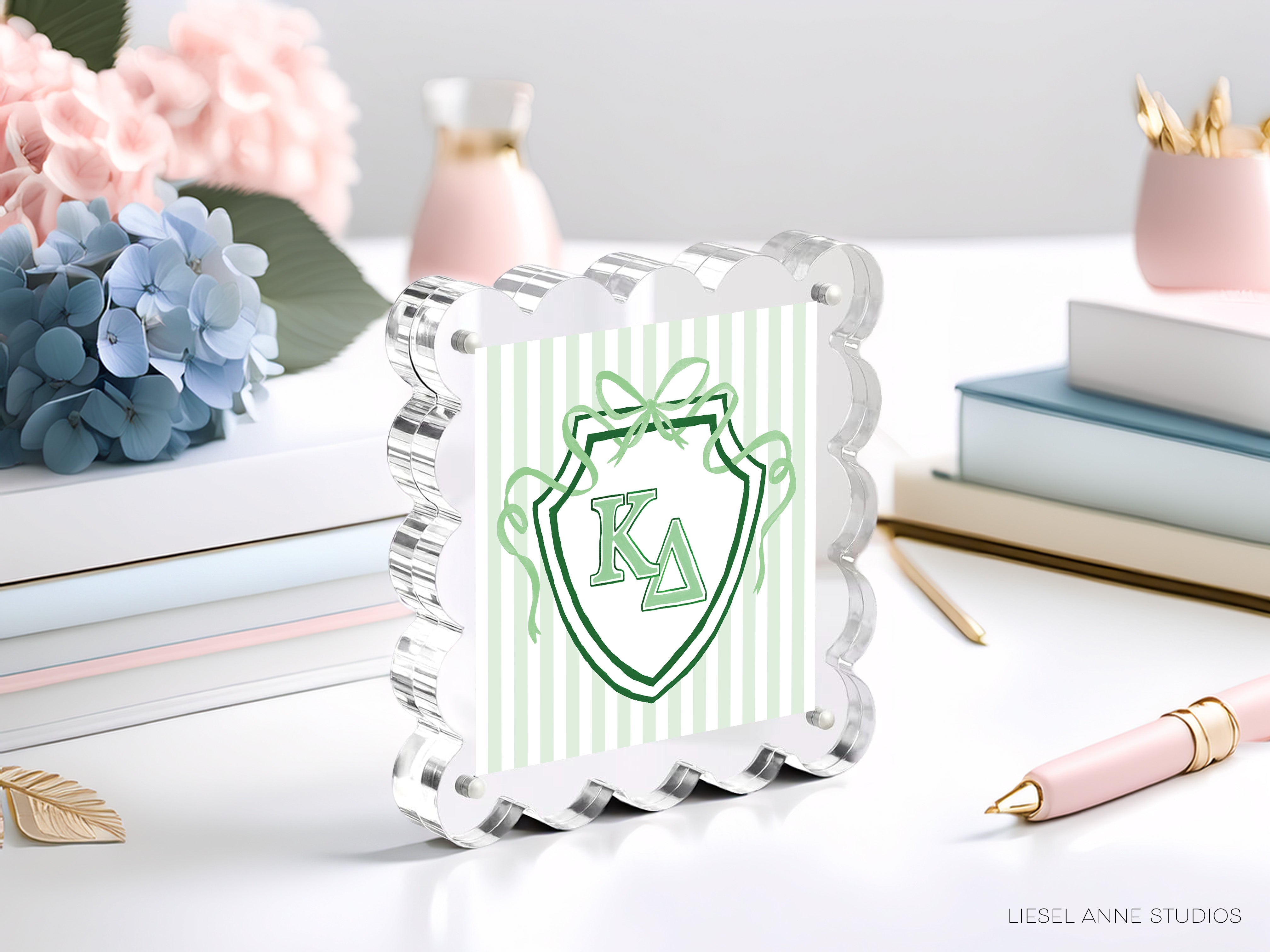 a clear glass frame with a decorative border, displaying a green and white shield-shaped logo with the letters "KA" in the center. The frame is placed on a table surrounded by various items, including a stack of books, a pink vase, and a pink pen.