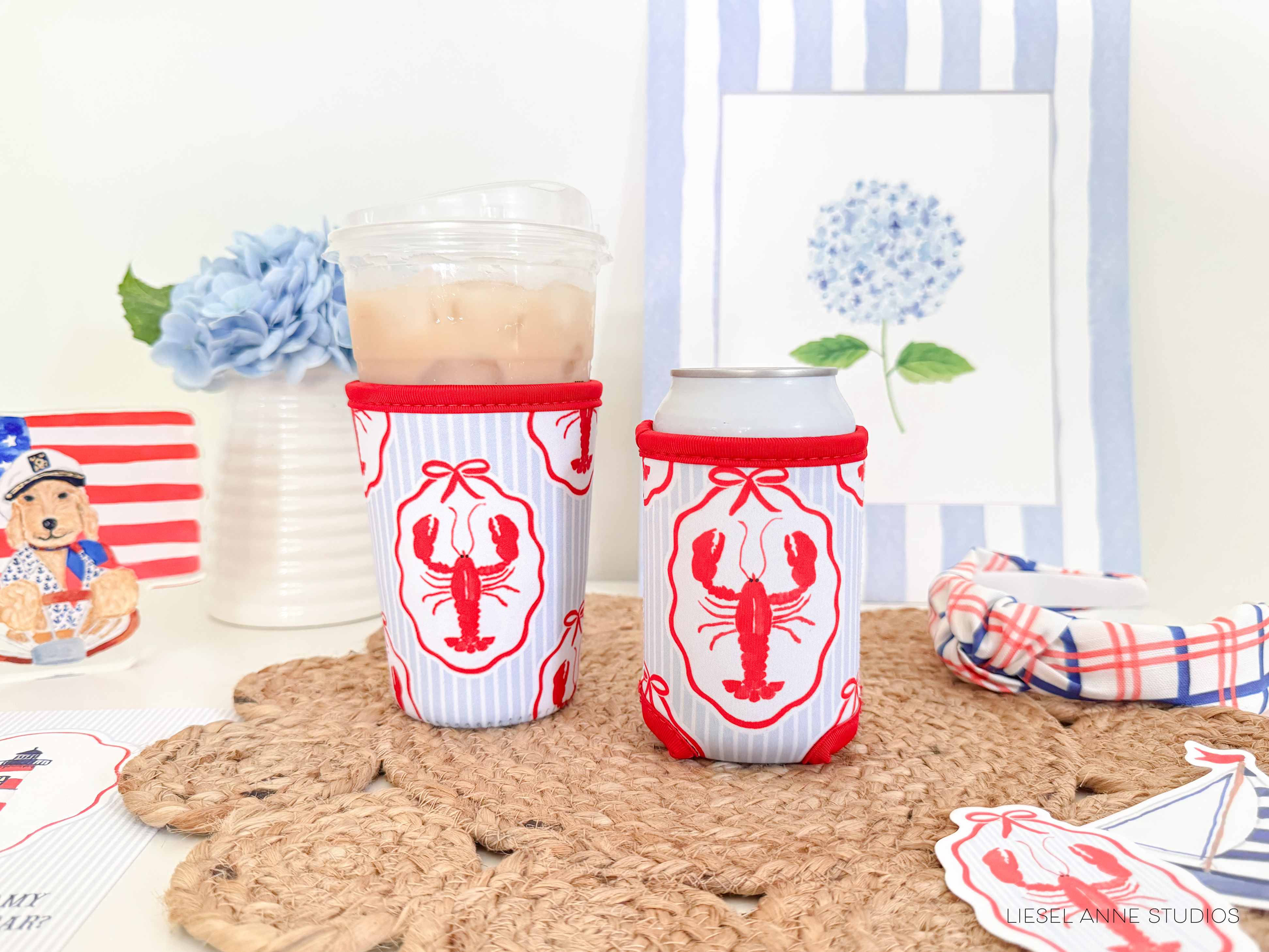 two red and white striped lobster-themed drink holders, each containing a beverage, placed on a woven placemat. In the background, there is a framed picture of a blue hydrangea flower, and a small American flag-themed decoration.