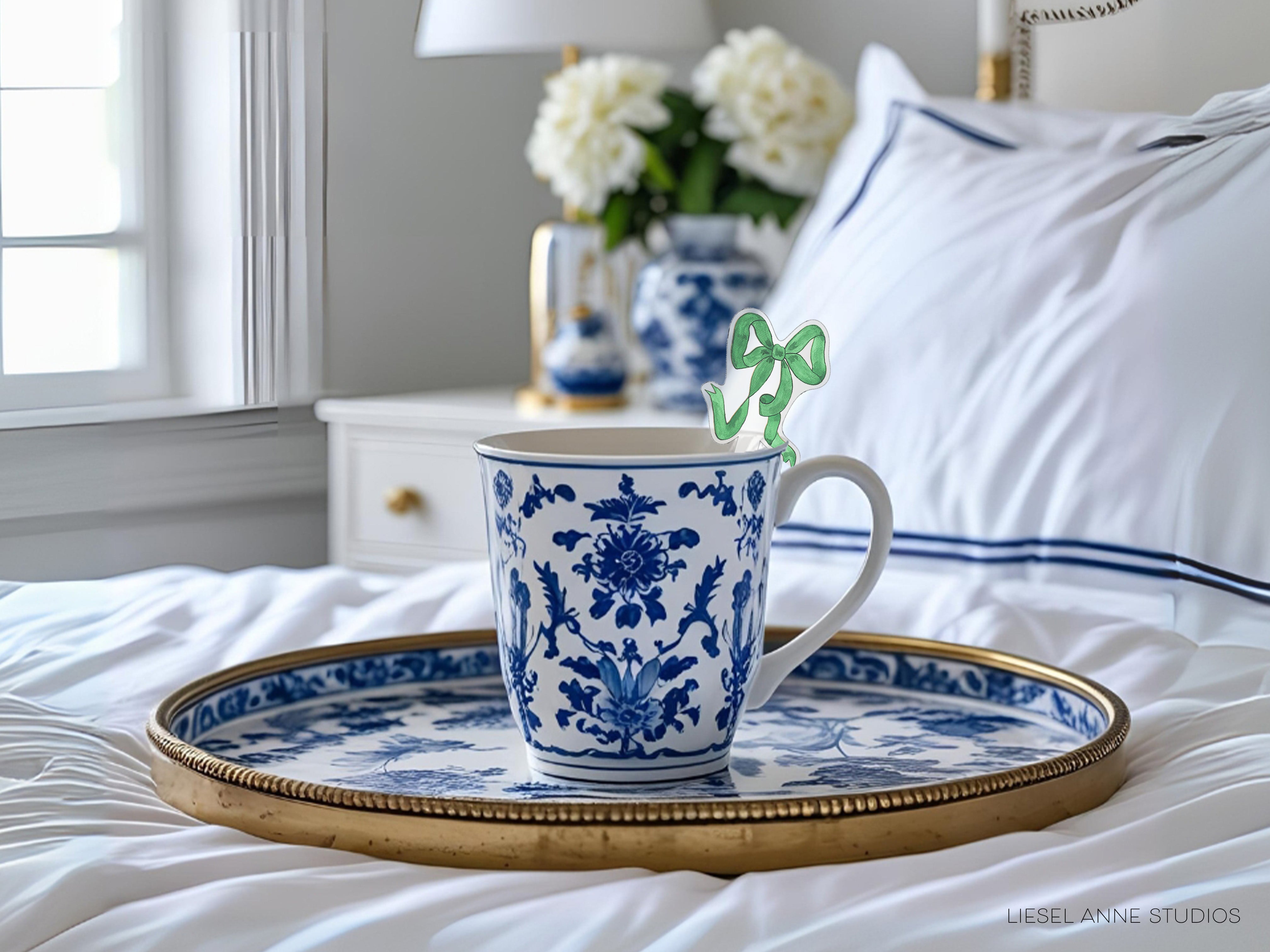 A blue and white patterned mug with a green bow sits on a gold tray, placed on a bed with white sheets and a white pillow.