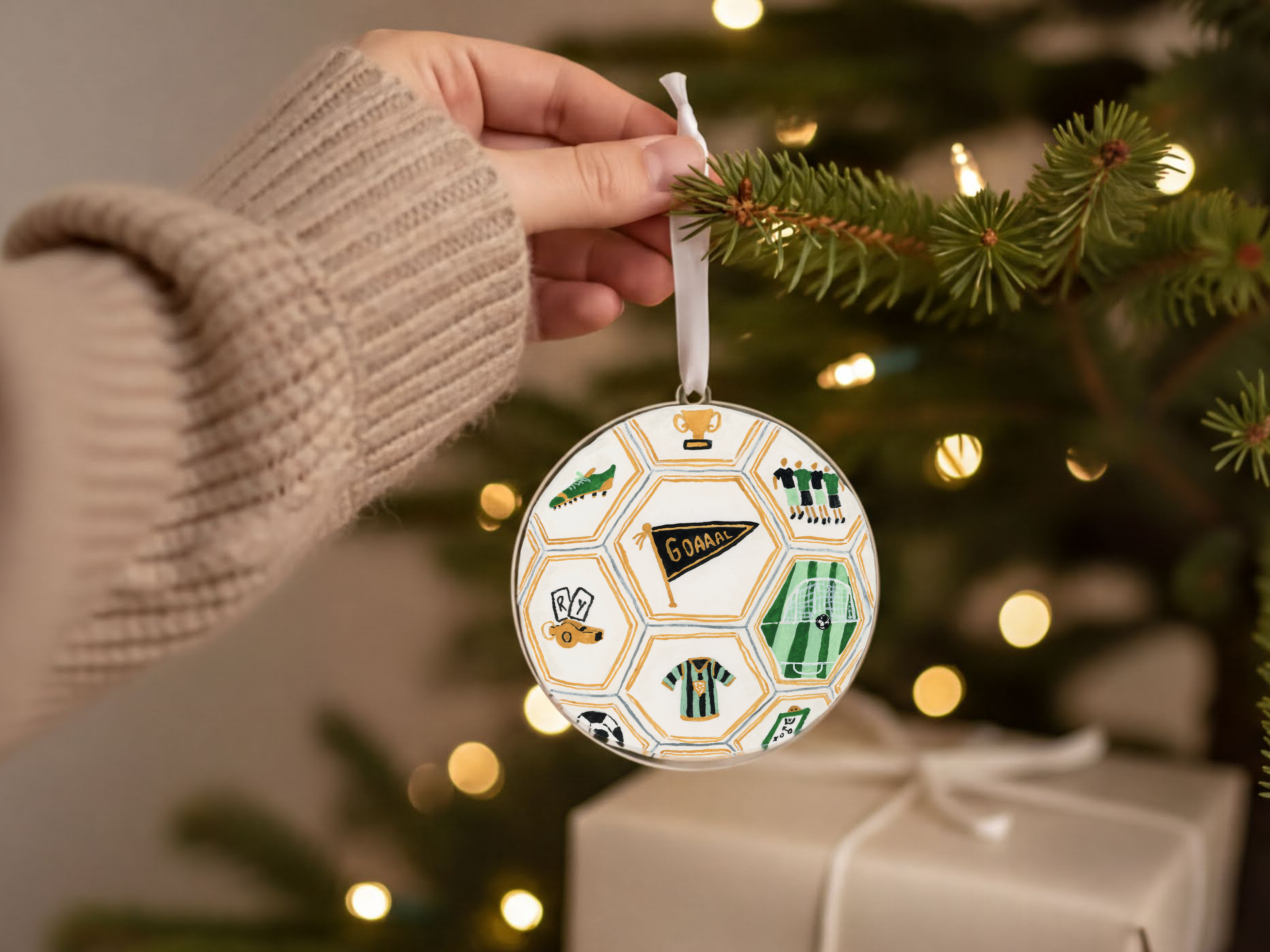 A hand holding a round ornament with soccer-themed illustrations, including a soccer ball, jersey, and trophy, hanging from a Christmas tree.