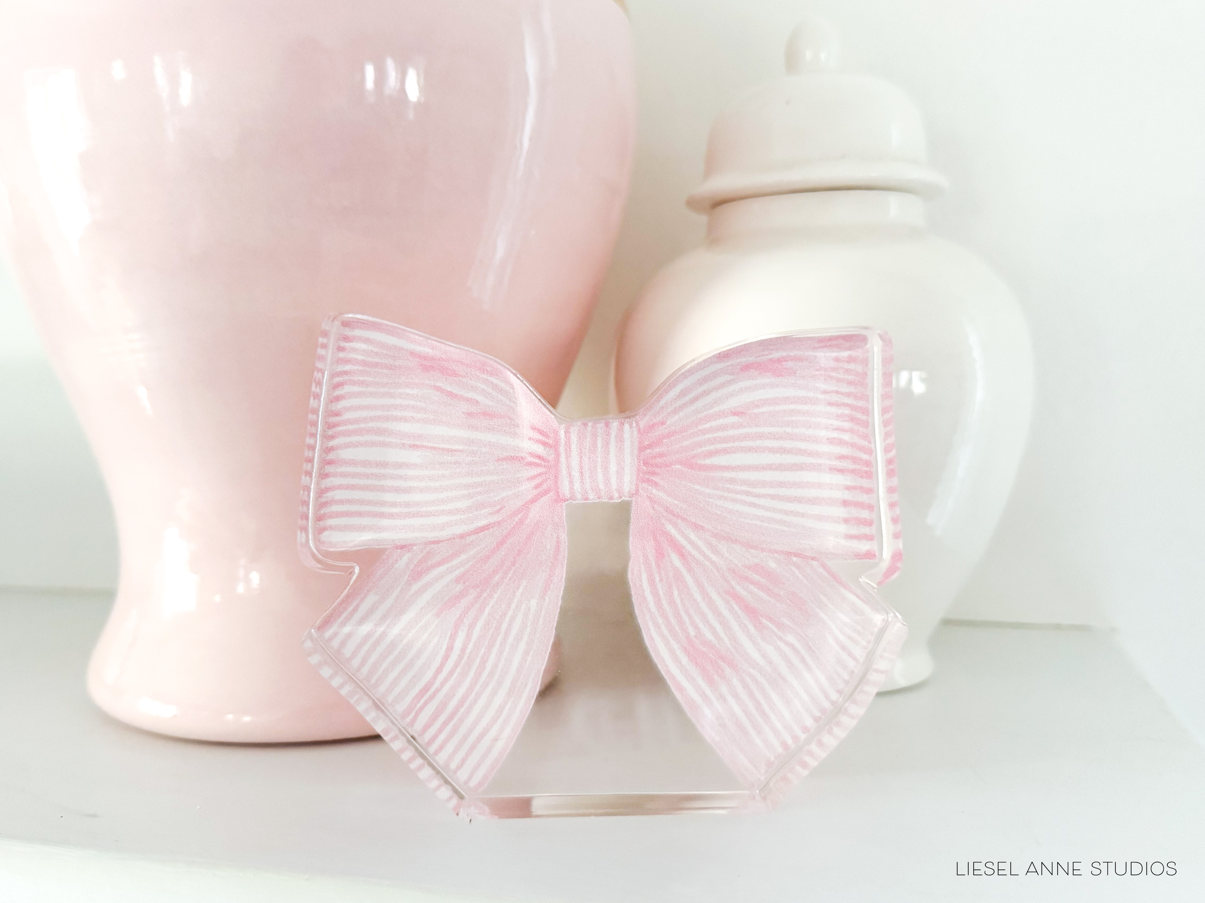 A pink bow tie is displayed in front of a white vase and a white ceramic jar on a white shelf.