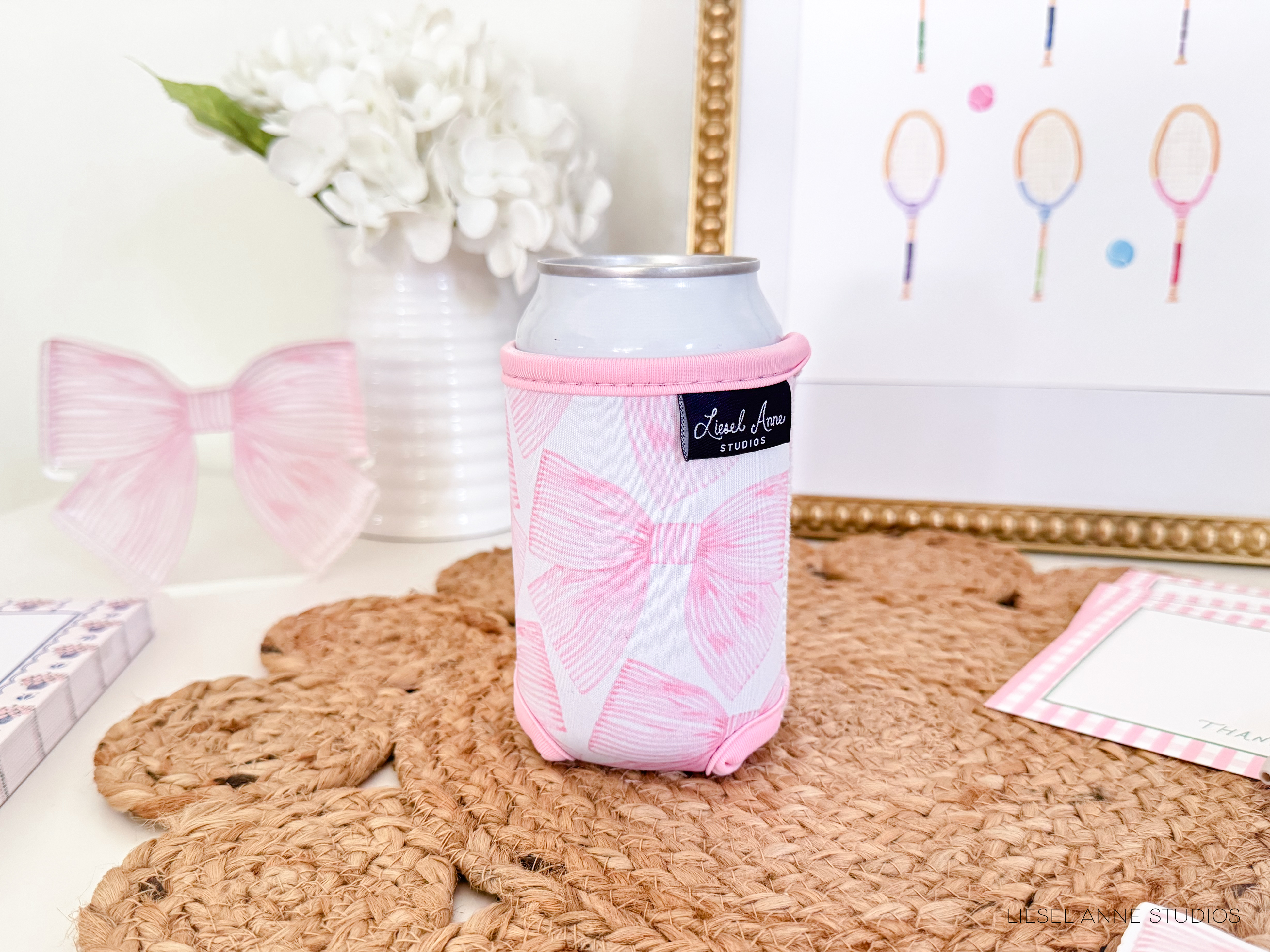 a pink and white bow-patterned can holder placed on a woven placemat, with a framed picture of tennis rackets in the background.