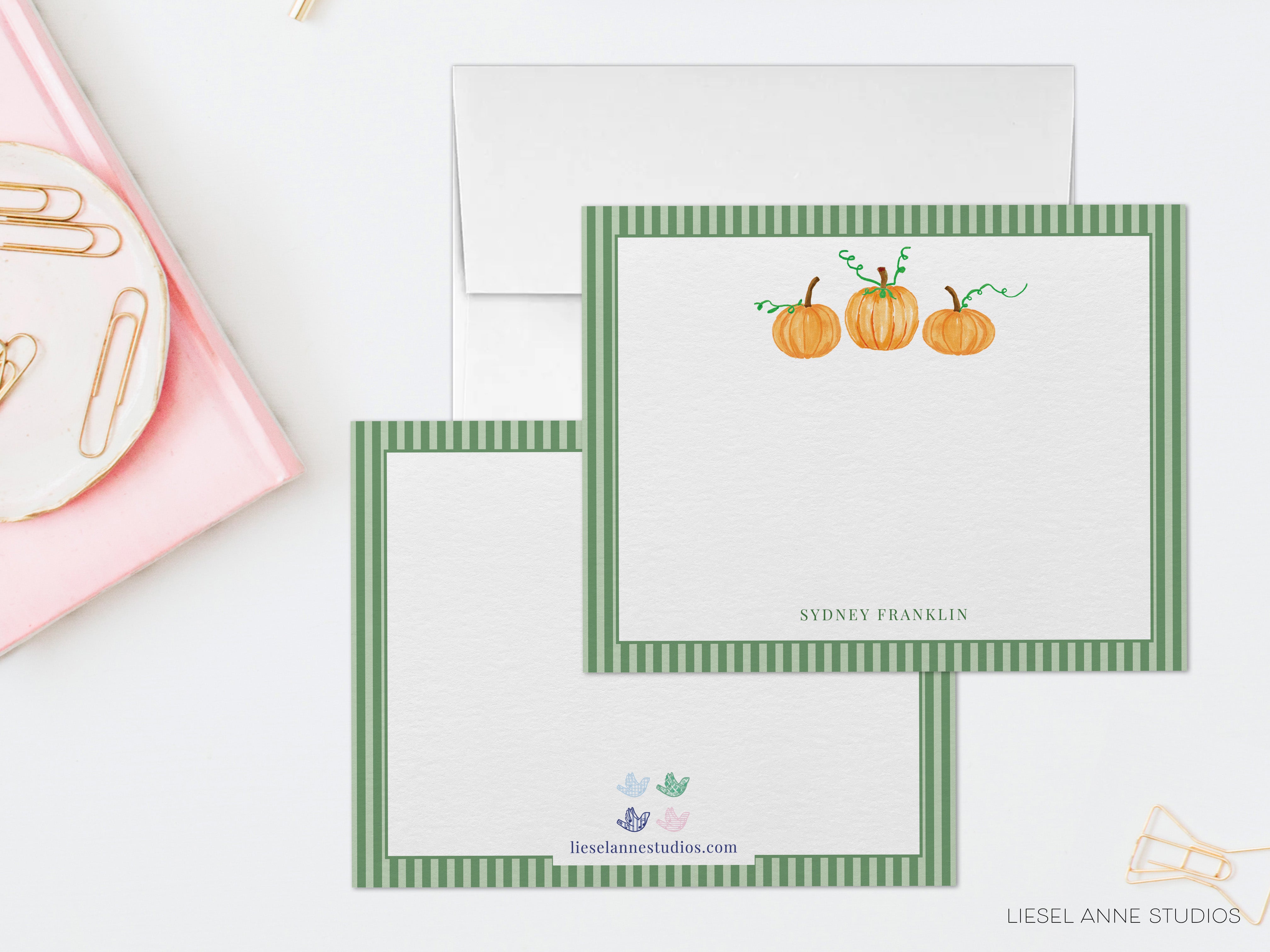 two blank greeting cards with a green and white striped border, each with a pumpkin illustration on the front. The cards are placed on a white surface, and there is a pink object in the background.