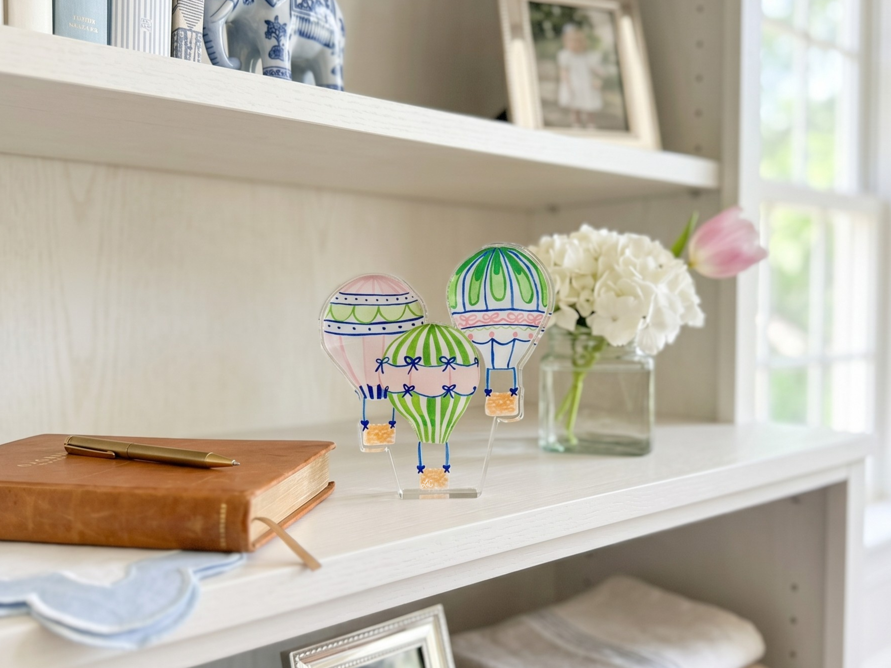 a white shelf with various decorative items, including two hot air balloons, a vase of white flowers, and a book.