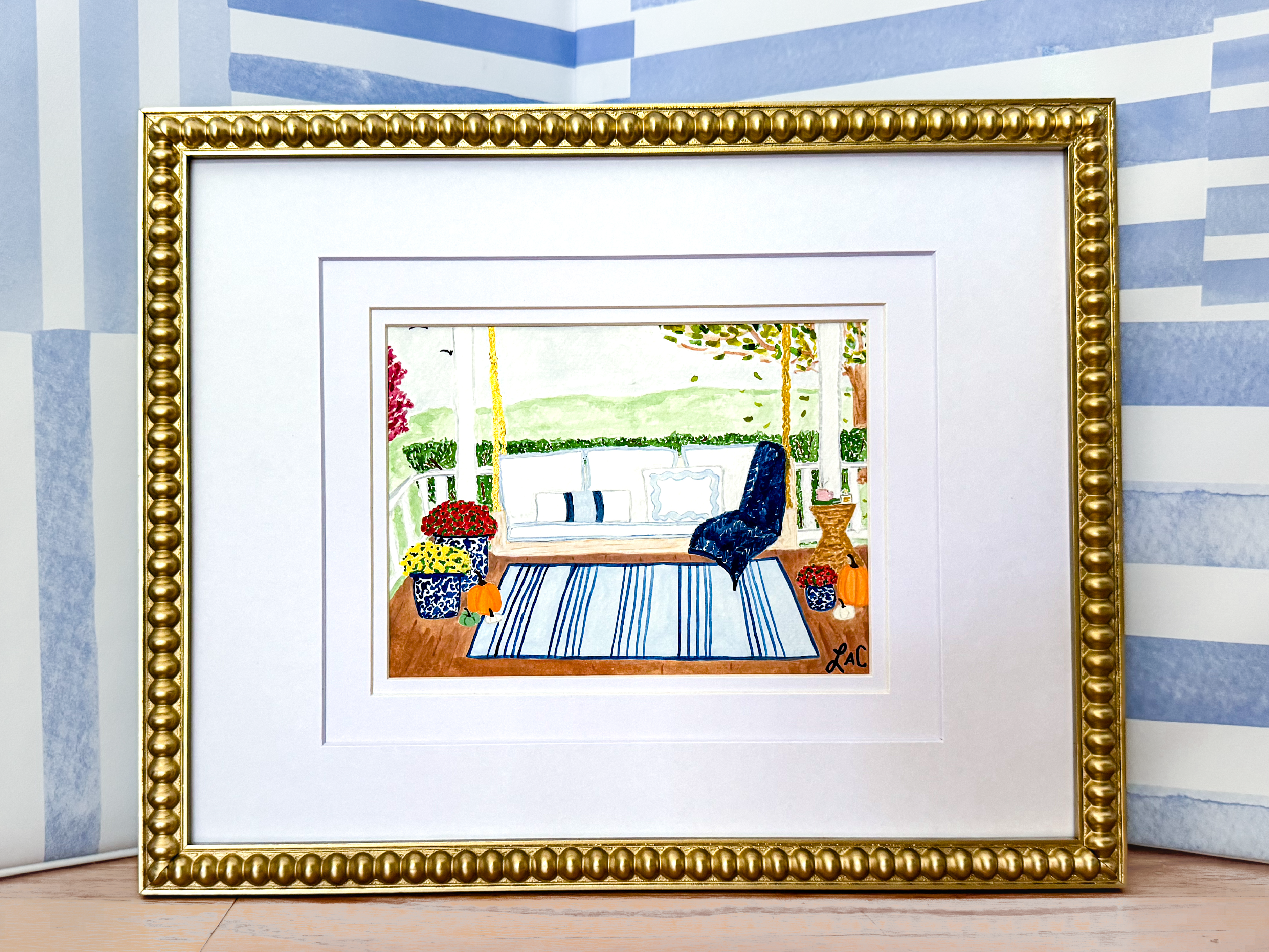a framed painting depicting a serene outdoor scene with a blue chair, a white bench, and a blue and white striped rug.