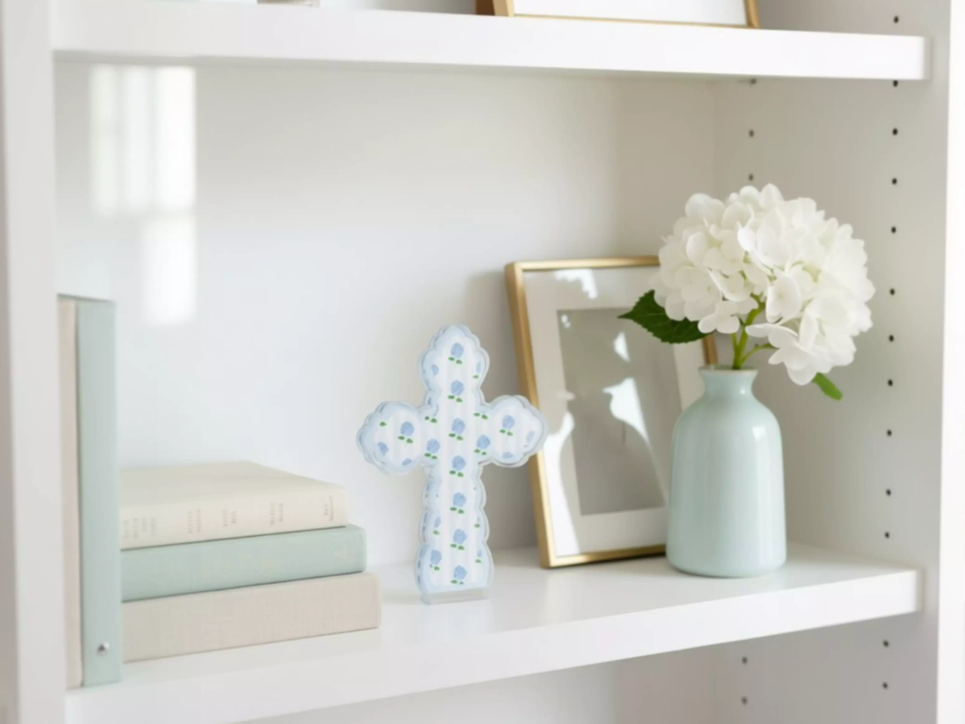 A white bookshelf with various items on it, including a blue and white cross, a white vase with white flowers, and a stack of books.