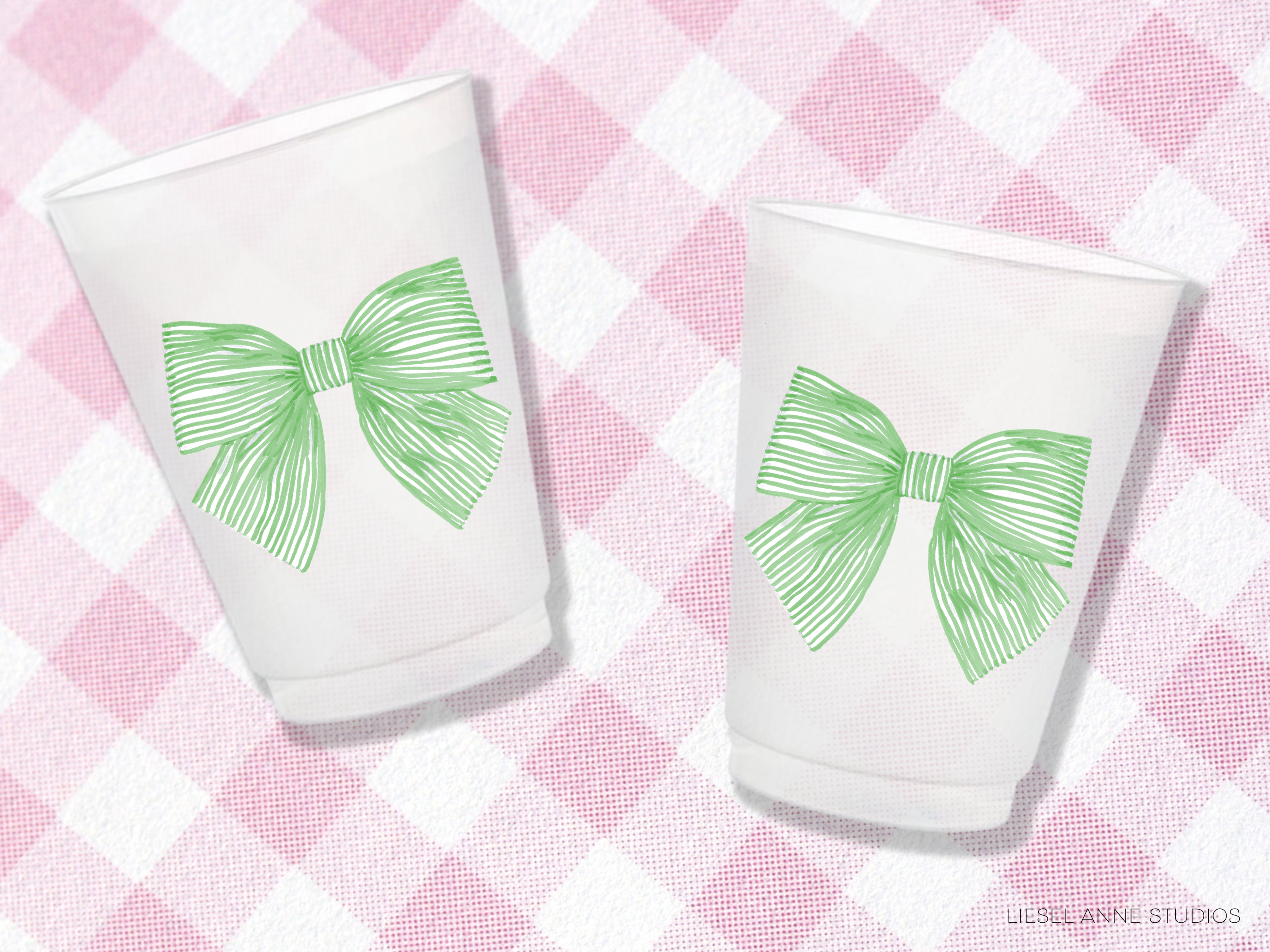 Two white cups with green bows on them, placed on a pink and white checkered background.