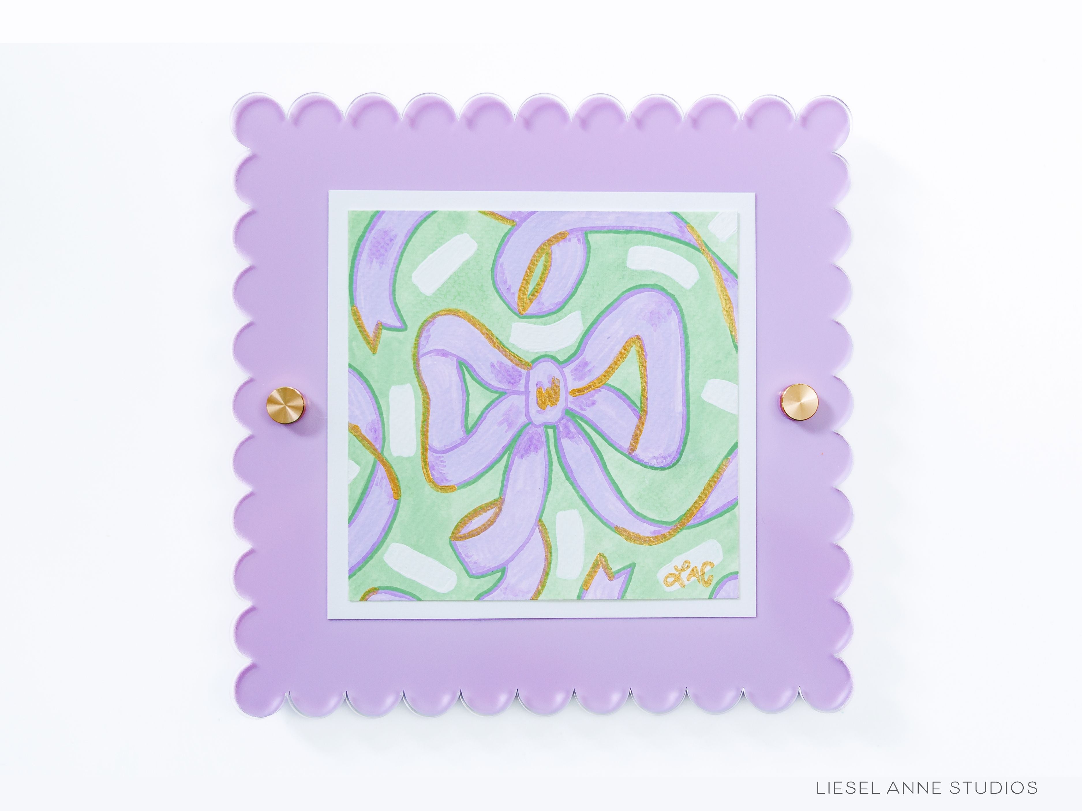 Golden Bows Green & Lavender 01 [Original Artwork + Lavender Scalloped Frame]-Original artwork measures 5"x5" | Hand-Painted with gouache on cold press watercolor paper | Matted and framed in an acrylic scalloped frame sized 8"x8" | Hanging hardware and acrylic clear stand included | Features our signature Golden Bows design with a light green base, lavender bows and shimmery gold accents | Initialed in gold by Liesel Anne (the artist) on the front and signed and dated on the back-The Singing Little Bird