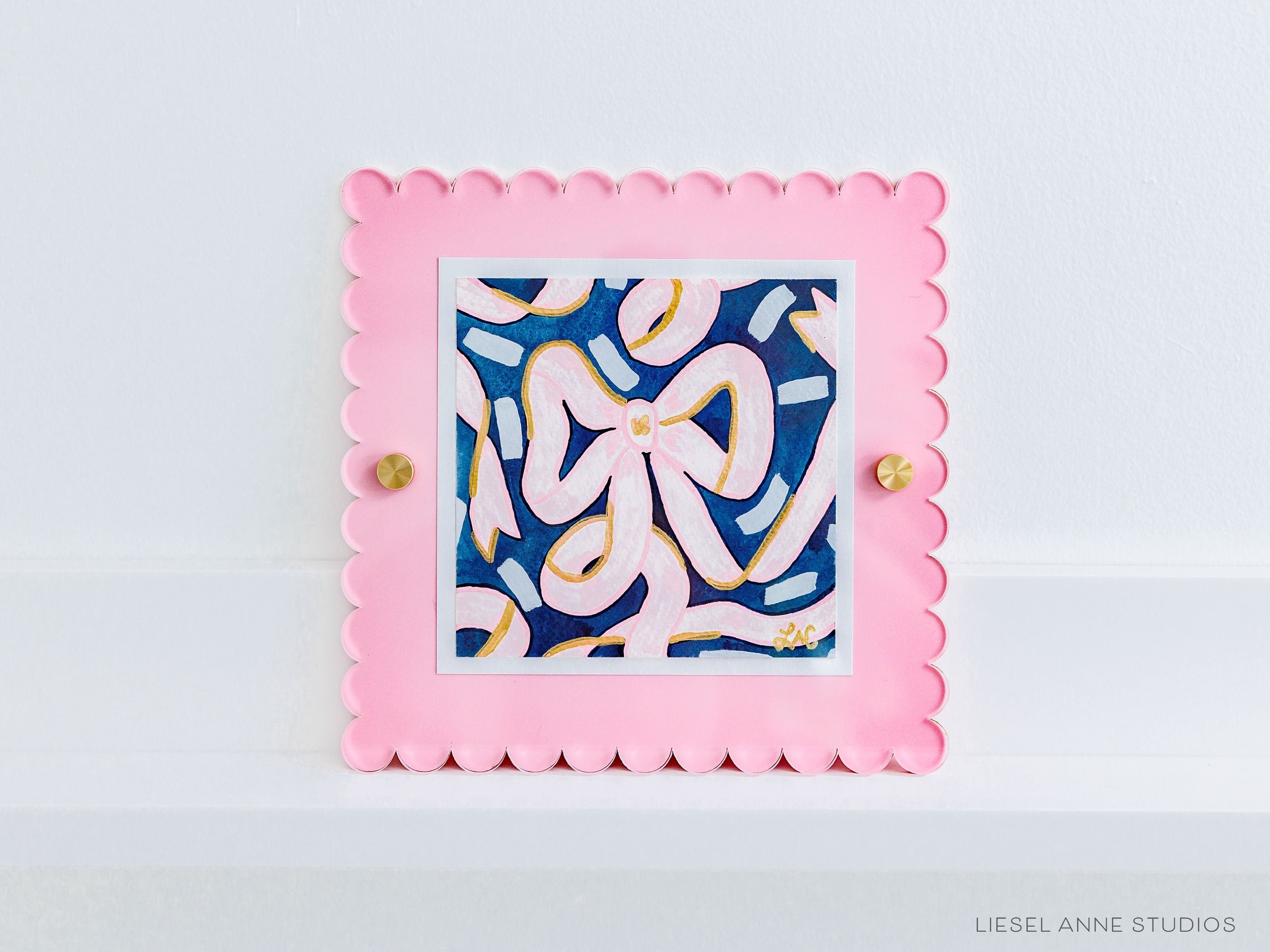 Golden Bows Navy & Pink 02 [Original Artwork + Pink Scalloped Frame]-Original artwork measures 5"x5" | Hand-Painted with gouache on cold press watercolor paper | Matted and framed in an acrylic scalloped frame sized 8"x8" | Hanging hardware and acrylic clear stand included | Features our signature Golden Bows design with a navy blue base, light pink bows and shimmery gold accents | Initialed in gold by Liesel Anne (the artist) on the front and signed and dated on the back-The Singing Little Bird