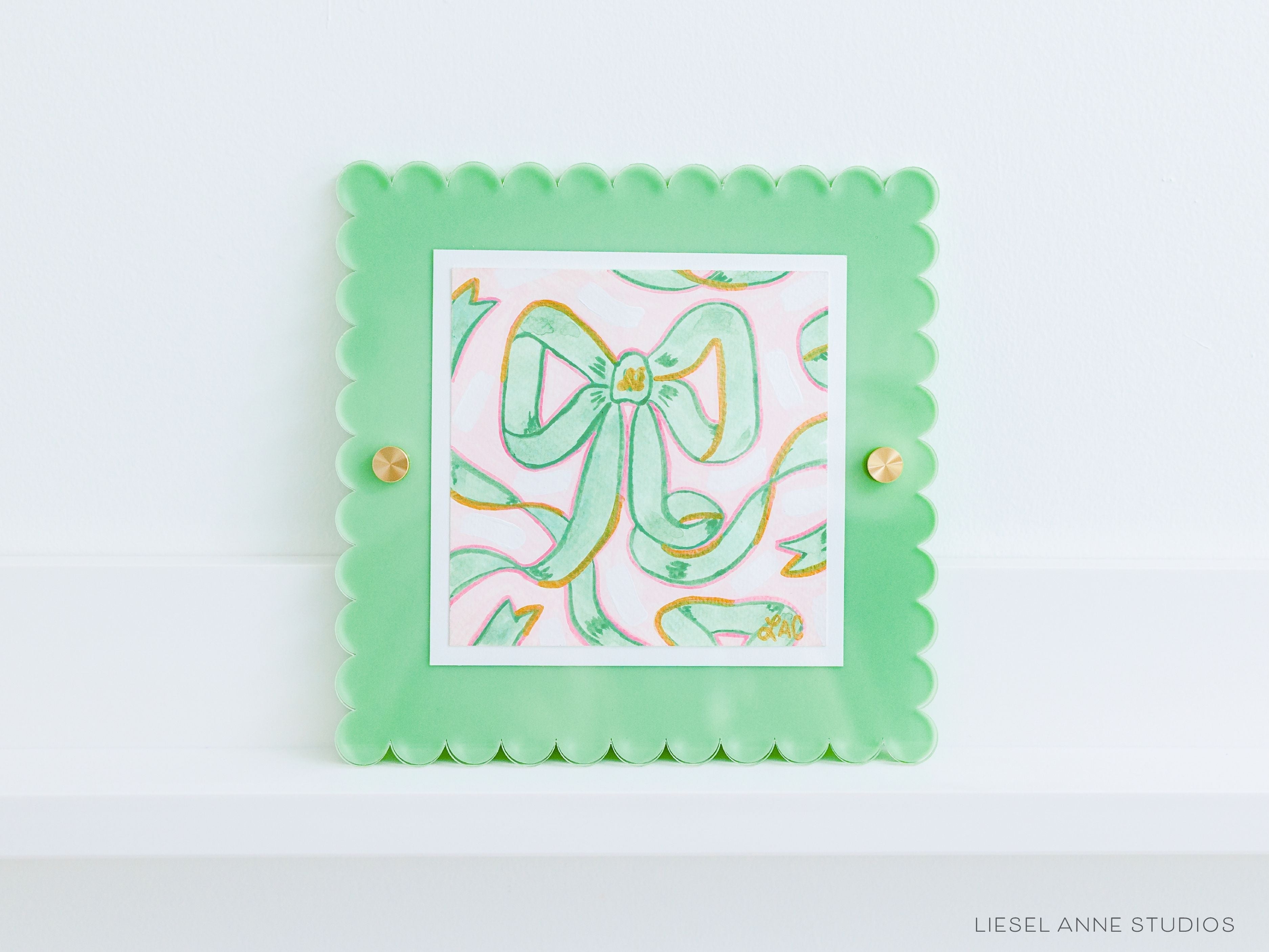 Golden Bows Pink & Green 03 [Original Artwork + Green Scalloped Frame]-Original artwork measures 5"x5" | Hand-Painted with gouache on cold press watercolor paper | Matted and framed in an acrylic scalloped frame sized 8"x8" | Hanging hardware and acrylic clear stand included | Features our signature Golden Bows design with a light pink base, green bows and shimmery gold accents | Initialed in gold by Liesel Anne (the artist) on the front and signed and dated on the back-The Singing Little Bird