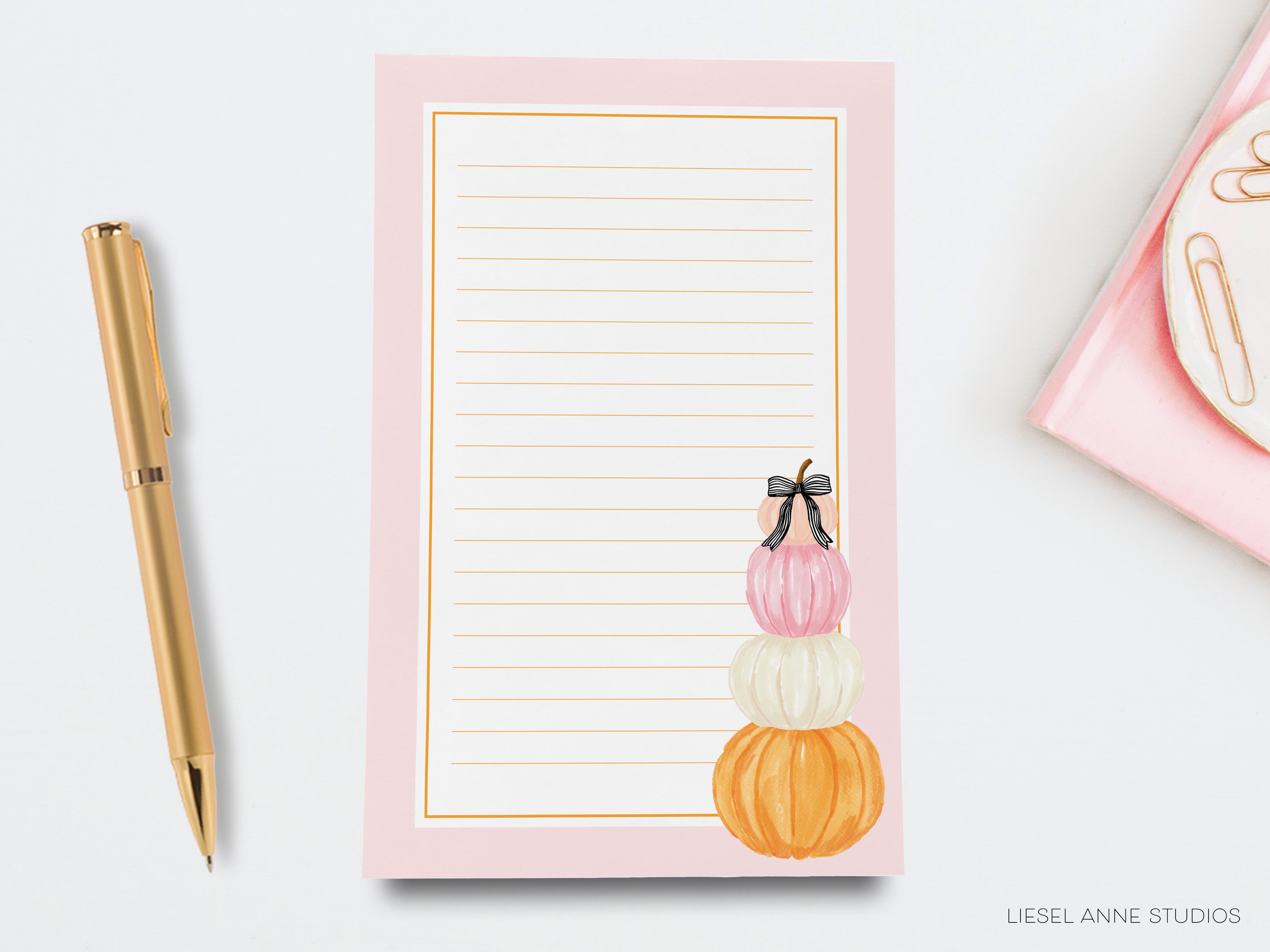 a pink lined paper pad with a decorative design of three stacked pumpkins, accompanied by a gold pen and a pink notebook on a white background.